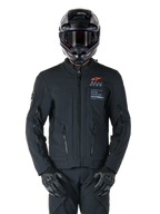 Alpinestars AMT-8 Stretch Drystar® XF Jacke, schwarze Motorradjacke, mit orangefarbenem Alpinestars-Logo und grauem AMT-Branding, hoher Kragen mit Frontreißverschluss und Belüftungsöffnungen, getragen mit schwarzem Helm und Handschuhen, schützende Adventure-Tourenbekleidung