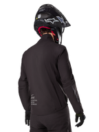 Alpinestars AMT-8 Stretch Drystar® XF Liner, schwarze wasserdichte Innenjacke, Rückansicht eines Fahrers mit schwarzem Helm und Handschuhen, mit Alpinestars Motion Technology-Branding am unteren Rücken, minimalistisches und funktionales Design für Adventure-Fahrten.