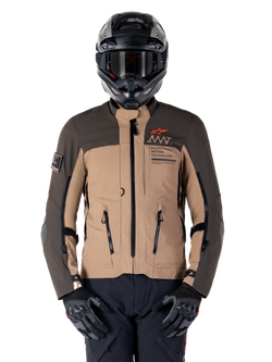 Alpinestars AMT-8 Stretch Drystar XF Jacke, Motorrad-Adventure-Touringjacke, Dunkelkhaki und Dunkelbraun, mit orangefarbenem Astars-Logo und schwarzen Reißverschlüssen, getragen mit schwarzem Adventure-Helm und Handschuhen, komplette Fahrausrüstung