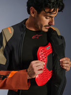 Alpinestars Halo Pro Drystar® XF gelamineerde jas in Walnut Rust Black, lifestyle-opname van een mannelijk model die de adventure-jas openritst om een rode borstprotector te onthullen, met oranje branding op de schouder en een zwarte basislaag met een rood logo.