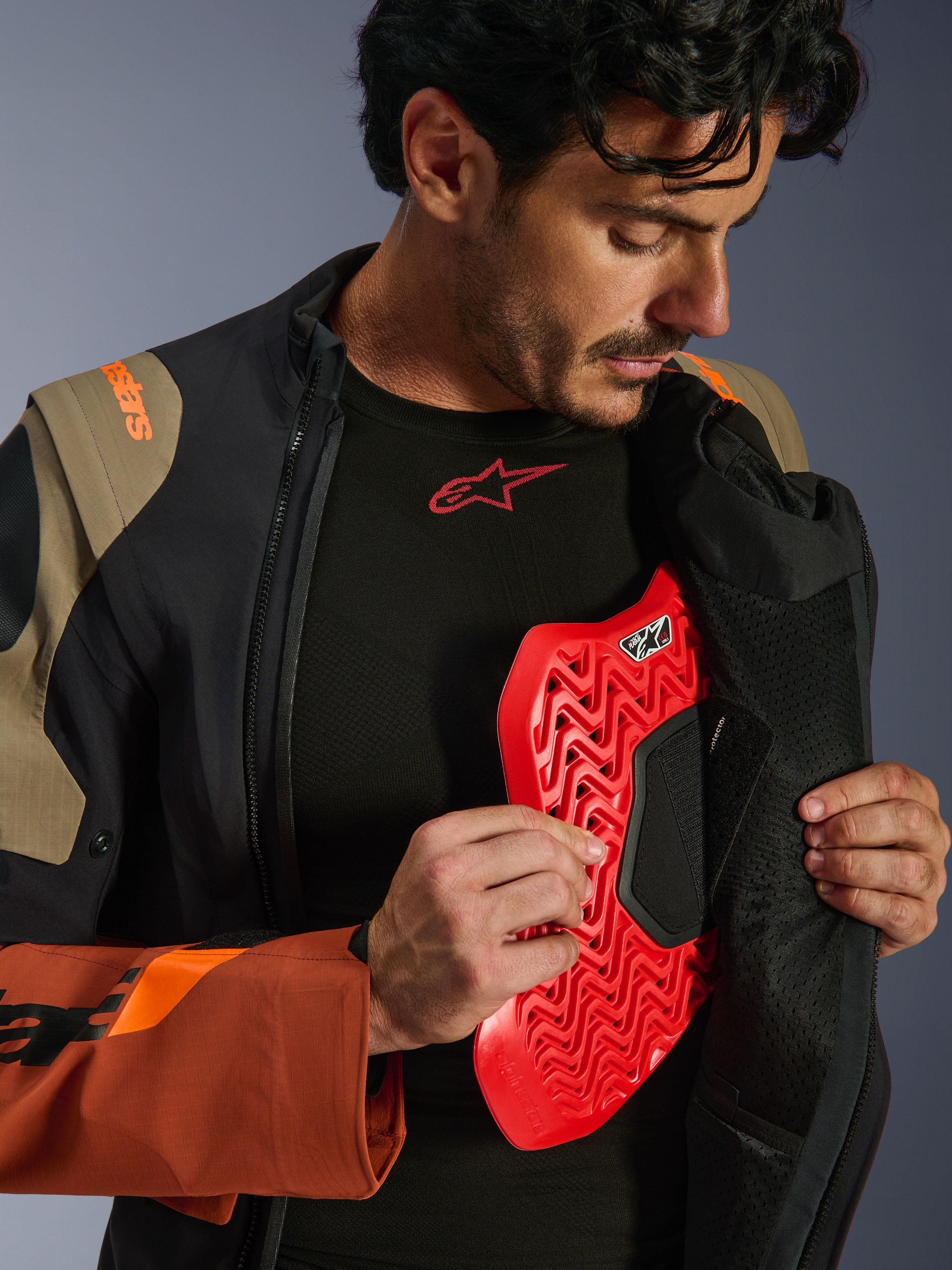 Alpinestars Halo Pro Drystar® XF gelamineerde jas in Walnut Rust Black, lifestyle-opname van een mannelijk model die de adventure-jas openritst om een rode borstprotector te onthullen, met oranje branding op de schouder en een zwarte basislaag met een rood logo.