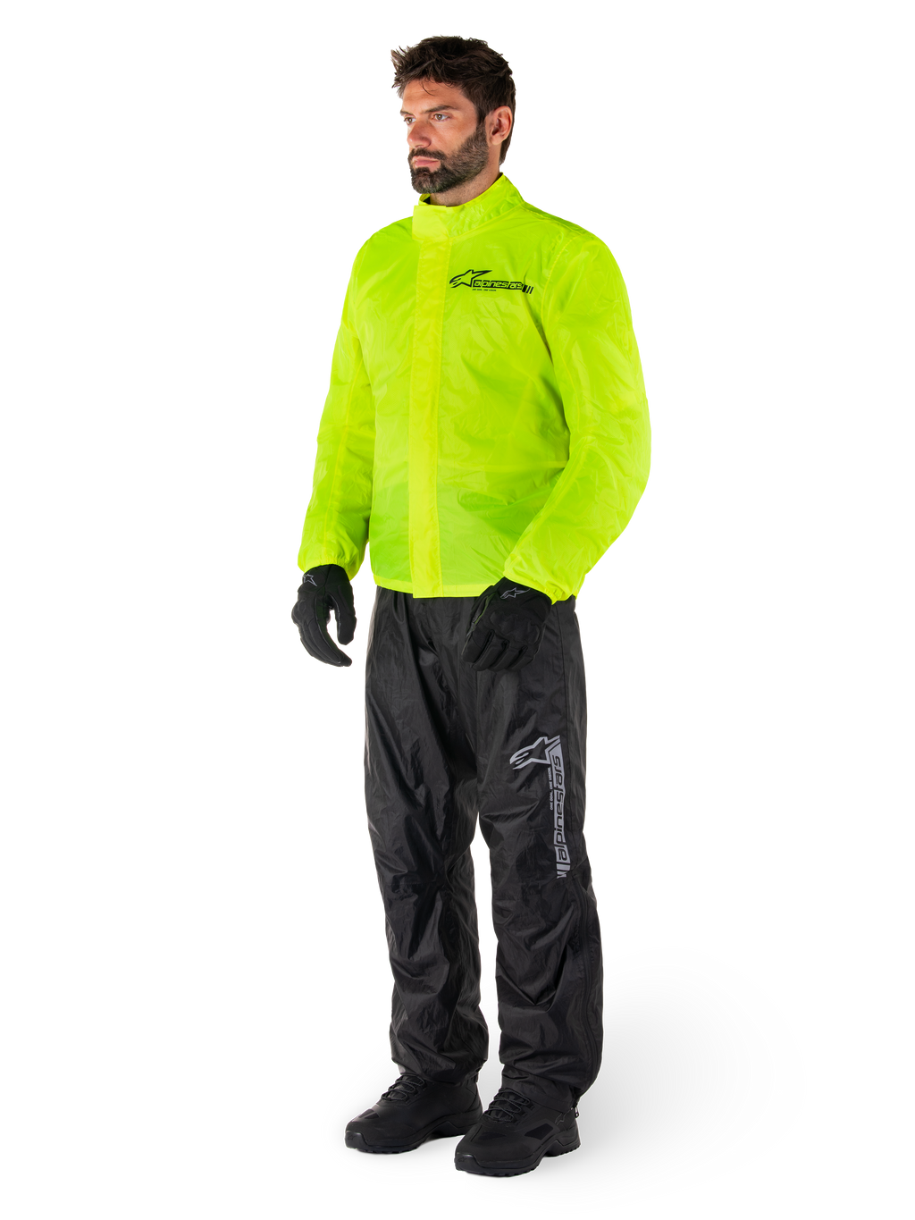 Homme portant la veste Alpinestars Hurricane Rain V2 en jaune fluo et le pantalon Hurricane Rain V2 en noir, équipement de pluie moto imperméable haute visibilité, logos réfléchissants sur la poitrine et la cuisse, tenue complète avec gants et bottes noirs