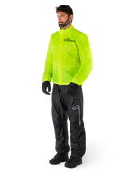 Homme portant la veste Alpinestars Hurricane Rain V2 en jaune fluo et le pantalon Hurricane Rain V2 en noir, équipement de pluie moto imperméable haute visibilité, logos réfléchissants sur la poitrine et la cuisse, tenue complète avec gants et bottes noirs