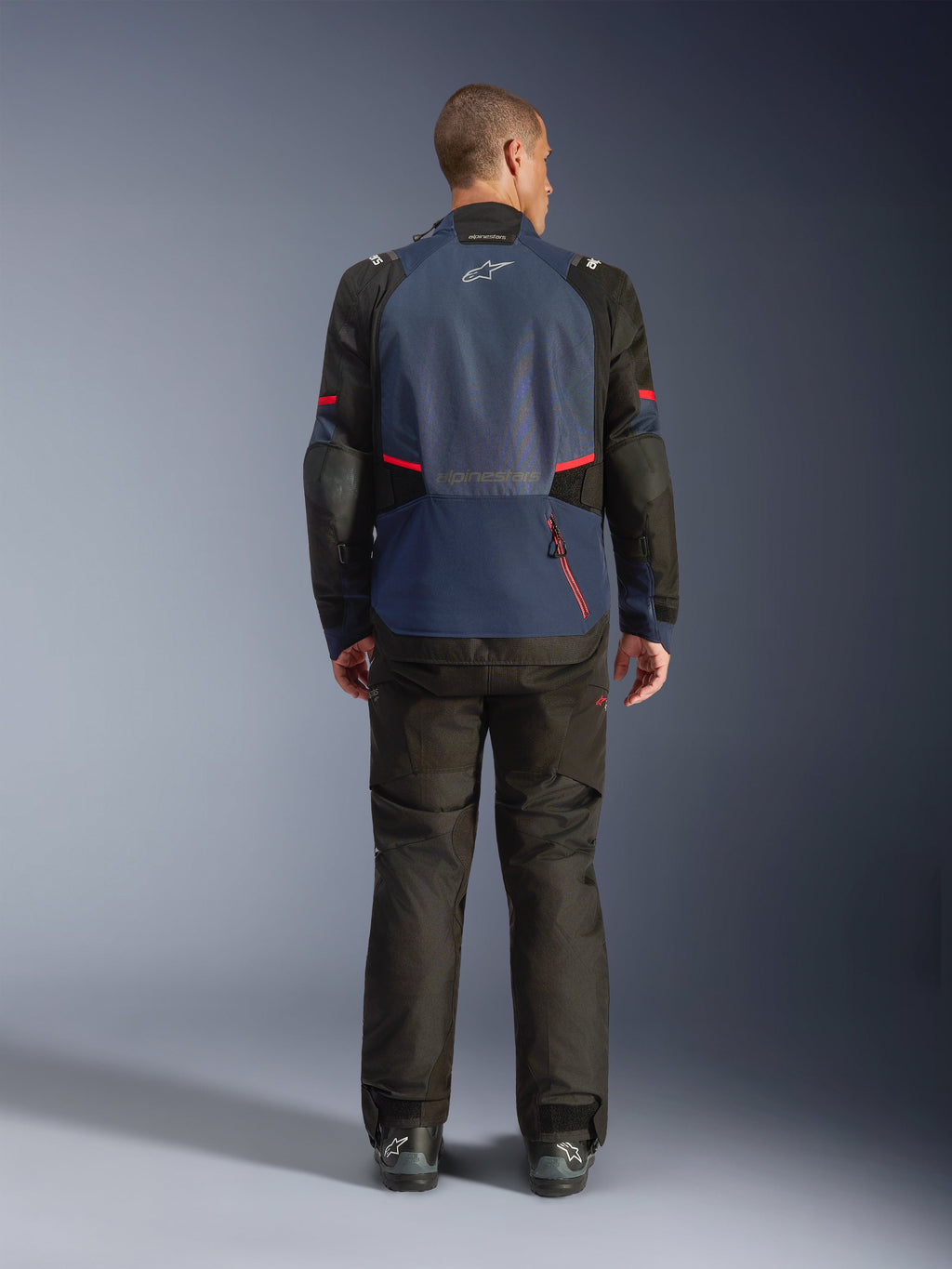 Alpinestars Andes V4 Drystar® Jacke, Rückansicht, Night Navy und Mood Indigo mit schwarzen Einsätzen und roten Akzenten, getragen mit passender schwarzer Hose, mit Alpinestars-Logo auf dem oberen Rücken und einem großen Reißverschluss für das Rückenfach.