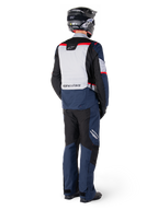 Vue arrière d'un pilote portant la veste imperméable Alpinestars ST-1 WP en bleu foncé, noir et rouge vif, associée à un pantalon imperméable assorti et un casque noir, présentant un panneau dorsal gris proéminent avec le logo Alpinestars et un design de tourisme d'aventure.