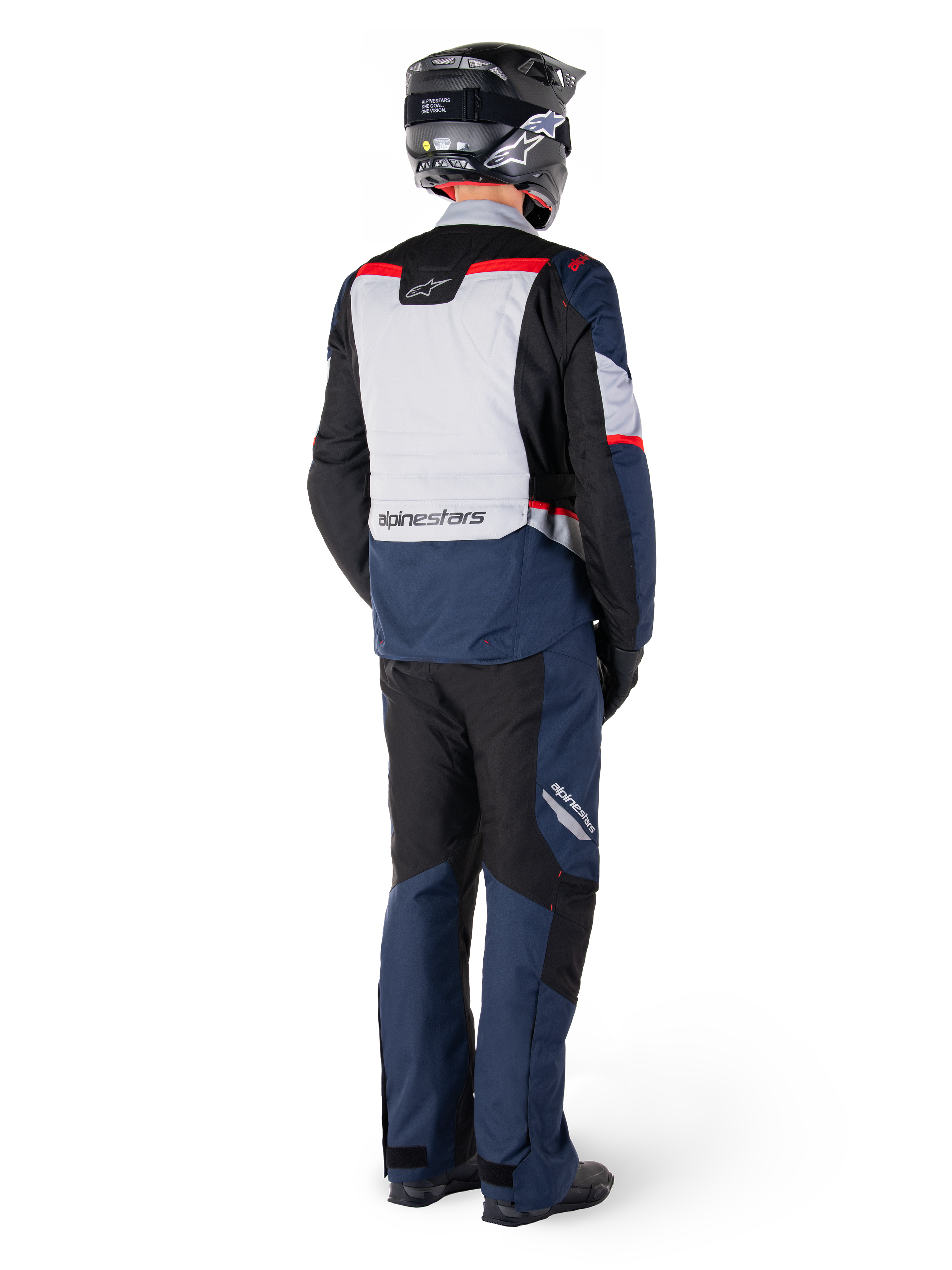 Vue arrière d'un pilote portant la veste imperméable Alpinestars ST-1 WP en bleu foncé, noir et rouge vif, associée à un pantalon imperméable assorti et un casque noir, présentant un panneau dorsal gris proéminent avec le logo Alpinestars et un design de tourisme d'aventure.
