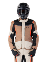 Alpinestars ST-1 Waterproof jas, Dark Khaki Sand Black Red Fluo, gedragen door een motorrijder met een zwarte helm en handschoenen, adventure touring motorkleding met beige en bruine panelen en felle fluo rode accenten, vooraanzicht op een witte achtergrond.