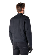 Alpinestars Clayton WR jas in het zwart, driekwart achteraanzicht op een mannelijk model, urban motorjas met gewatteerde vulling op de schouders en ellebogen, technische stiklijnen en reflecterend logo op de kraag, gecombineerd met een zwarte broek en handschoenen.
