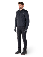 Mannelijke motorrijder die de Alpinestars Clayton WR jas draagt, zwarte urban motorjas, slim-fit ontwerp met gewatteerde schouderaccenten en matte afwerking, gecombineerd met een zwarte technische motorbroek en handschoenen.
