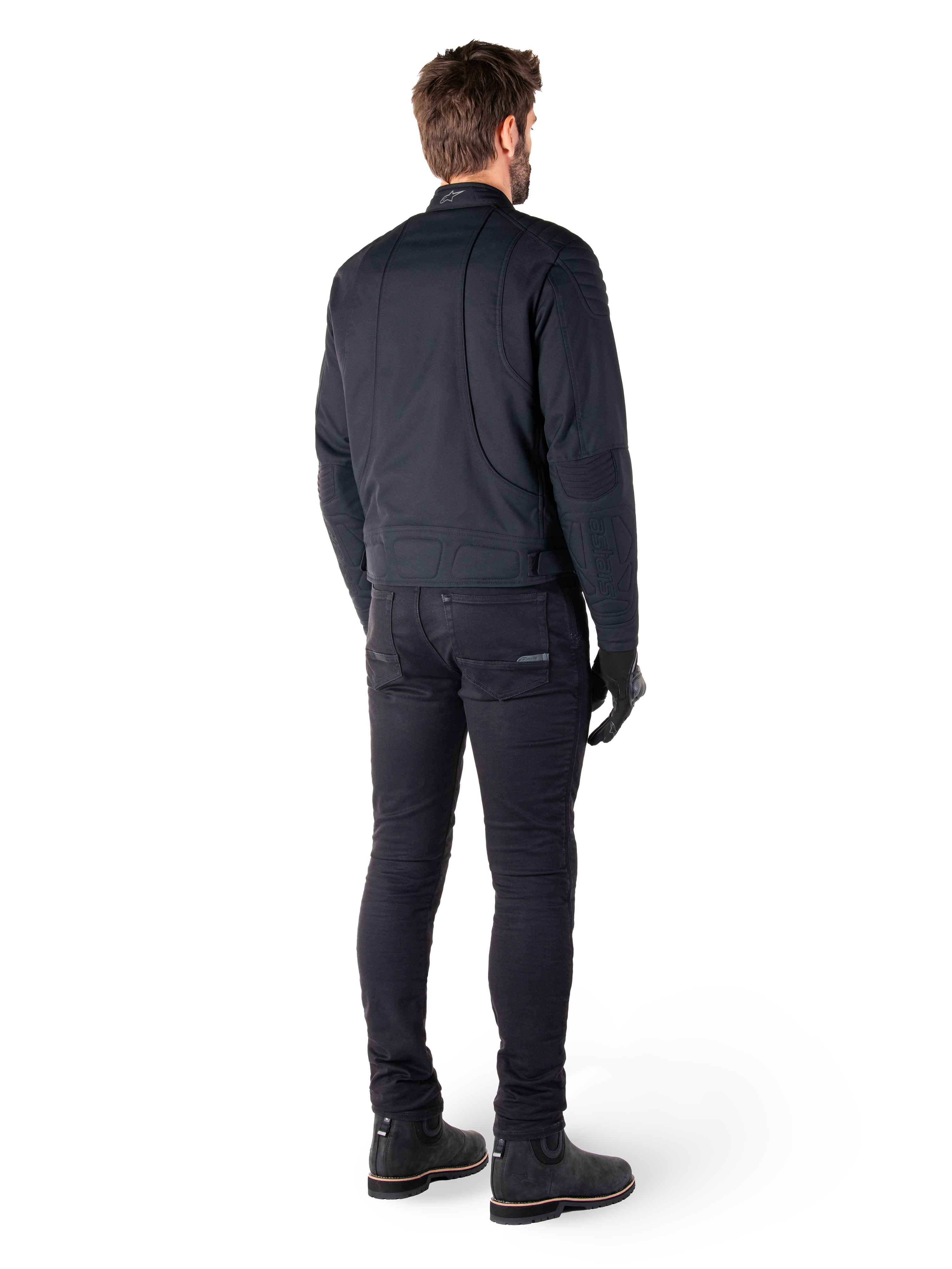 Achteraanzicht van een motorrijder die de Alpinestars Clayton WR Jas in het zwart draagt, voorzien van gewatteerde elleboog- en schouderpanelen, ergonomische gebogen stiknaden en reflecterende kraagdetails, gecombineerd met een zwarte motorbroek en laarzen voor een urban motorlook.