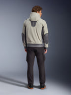 Alpinestars Moblast Waterproof Jacket, Iron Gray Dark Sage, rear view of a male rider wearing the hooded motorcycle jacket, light gray and dark sage colorway, vertical 'astars' logo on the upper back, dark gray elbow patches, styled with dark cargo pants for an urban contemporary look.