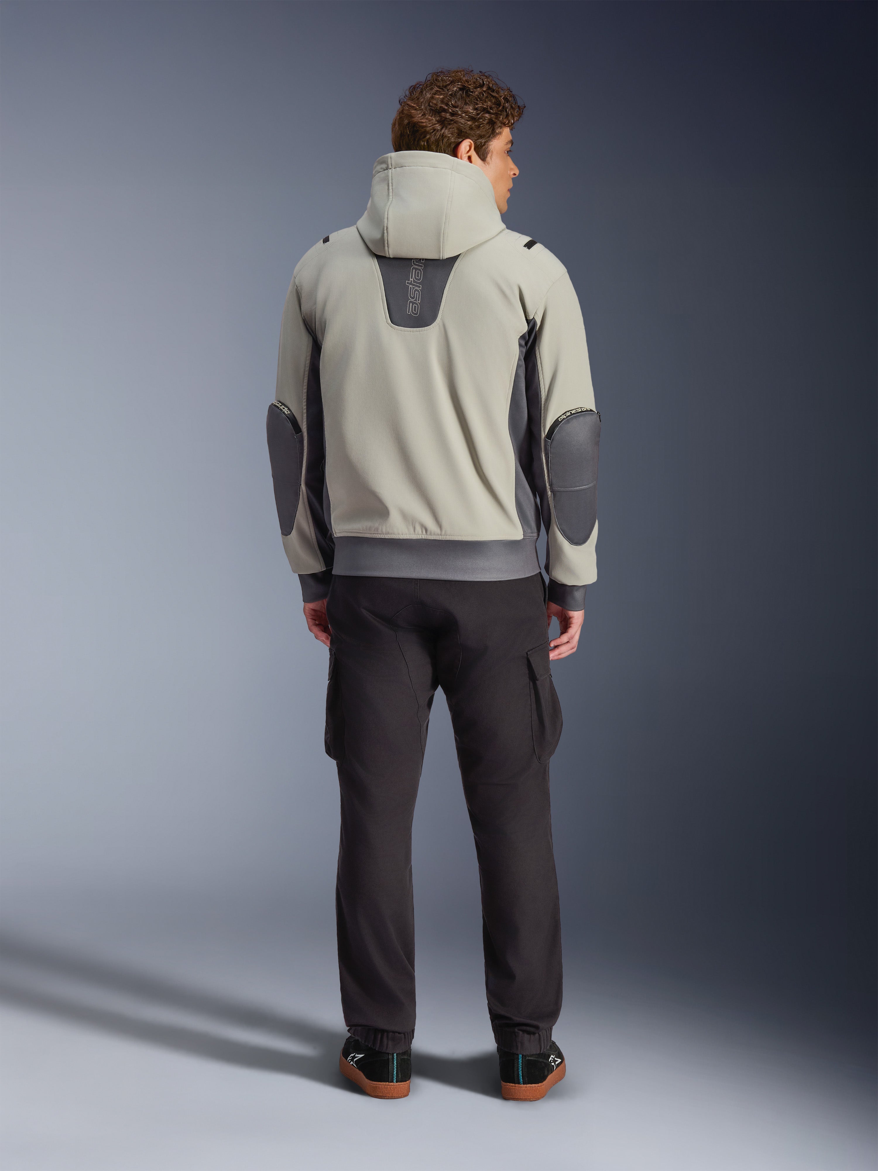 Alpinestars Moblast Waterproof Jacket, Iron Gray Dark Sage, rear view of a male rider wearing the hooded motorcycle jacket, light gray and dark sage colorway, vertical 'astars' logo on the upper back, dark gray elbow patches, styled with dark cargo pants for an urban contemporary look.