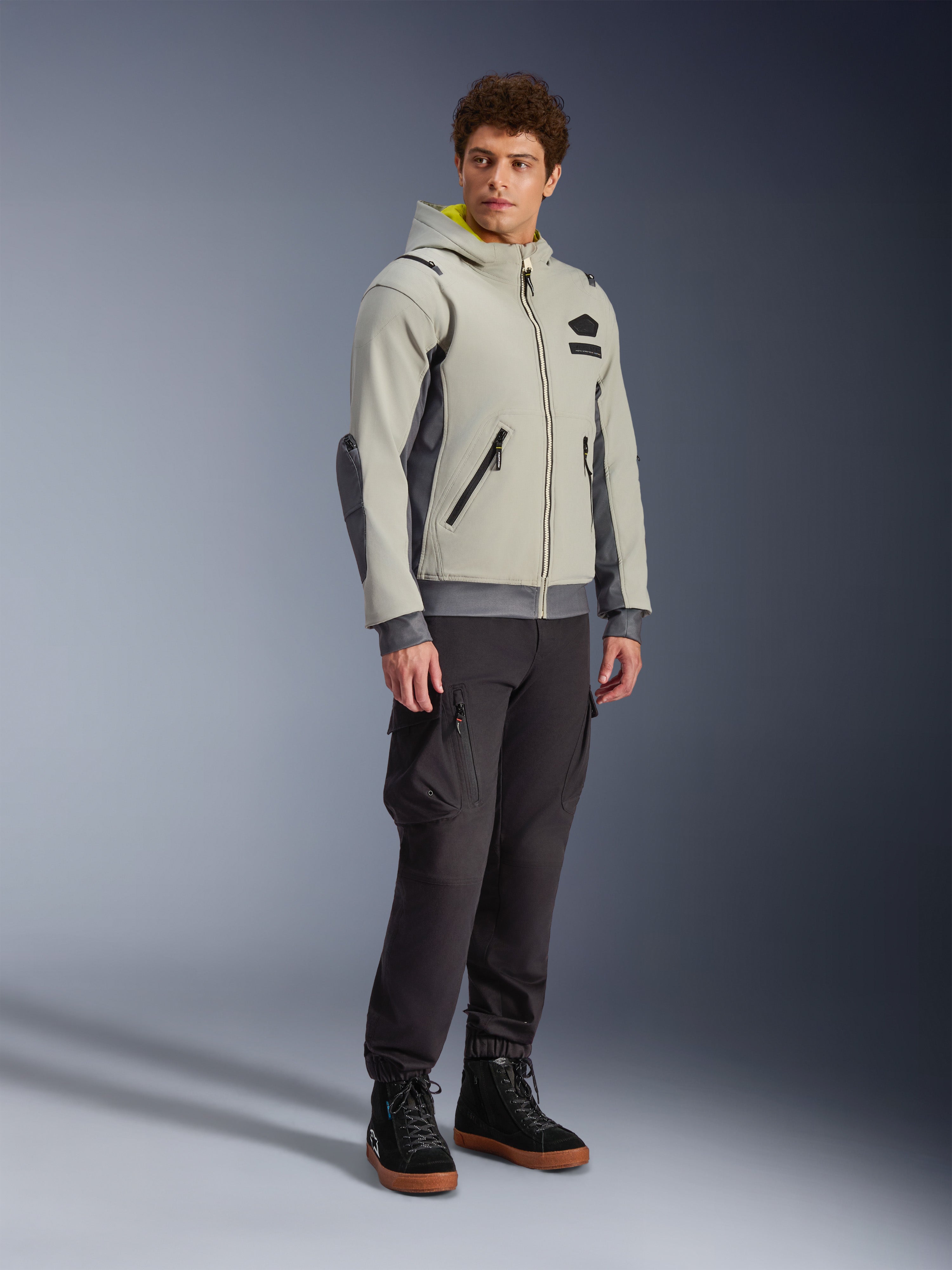 Alpinestars Moblast Waterproof Jacket, Iron Gray and Dark Sage, urban motorcycle riding jacket with a hoodie design, worn by a model with dark riding pants and black boots, minimalist cyber-tech aesthetic with a yellow interior hood and waterproof zippers