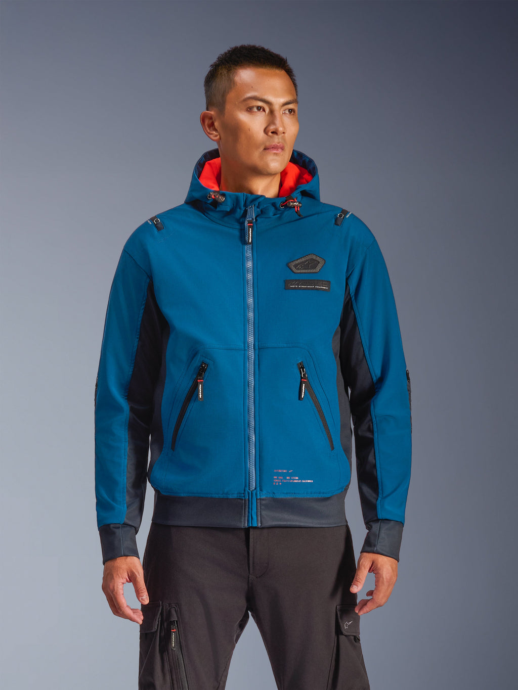 Alpinestars Moblast Waterproof Jacket gedragen door model, blauw en donkerblauw, urban motorjas, ontwerp met capuchon en rode binnenvoering, zwarte ritsen en patchdetail op de borst, op streetwear geïnspireerde motorkleding