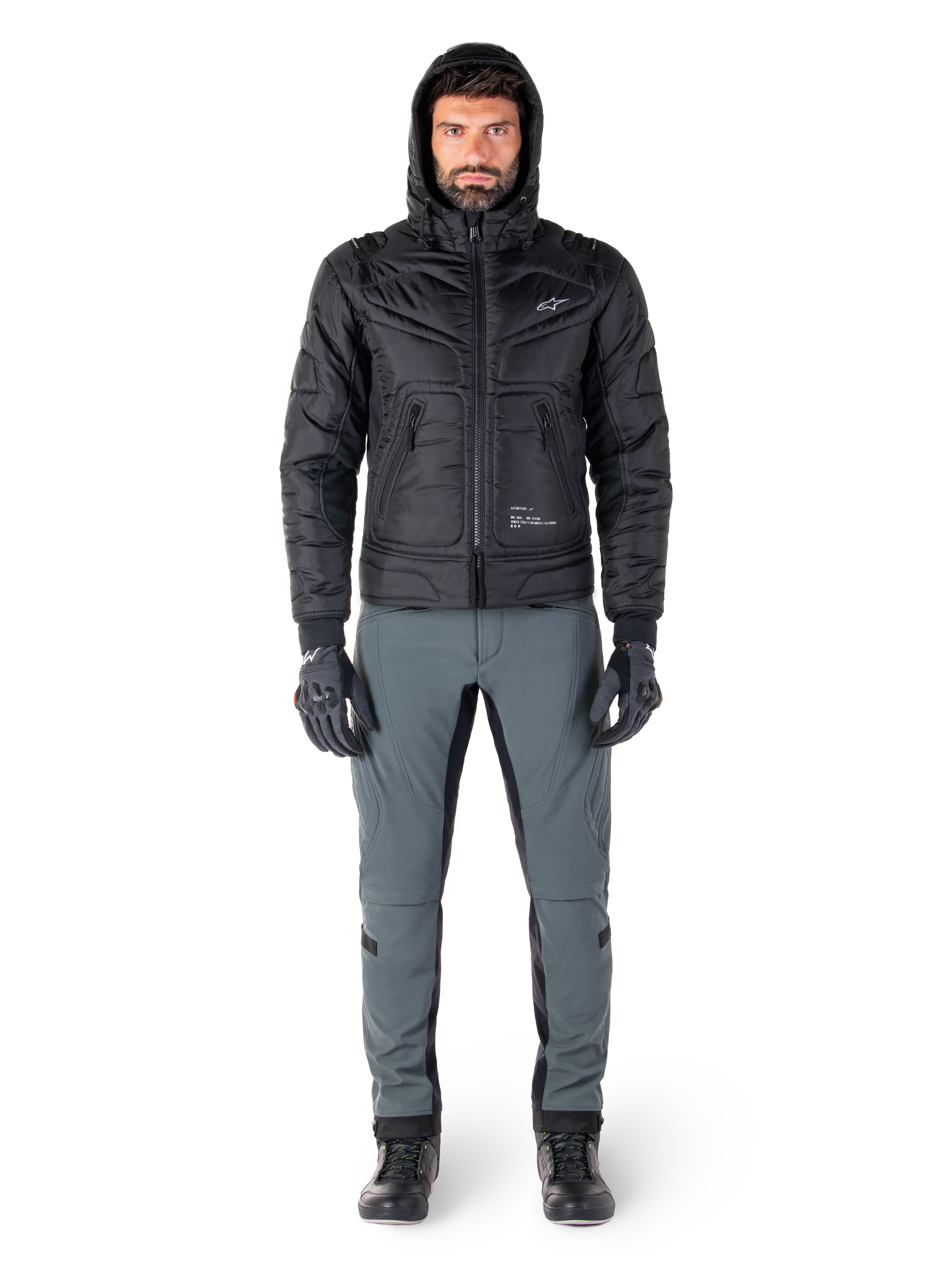 Alpinestars Mohobbs Waterproof Jas in zwart, mannelijk model draagt een gewatteerd bomberjack met capuchon, gecombineerd met een grijze motorbroek, zwarte handschoenen en zwarte laarzen, urban contemporary motoruitrusting
