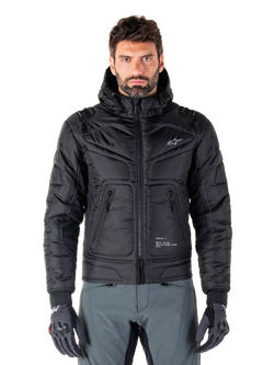 Alpinestars Mohobbs Waterproof Jacket, schwarze gepolsterte Bomber-Motorradjacke, gestepptes Polygewebe mit Frontreißverschluss und Reißverschlusstaschen, kleines weißes Logo auf der Brust, Modell trägt graue Hose und Handschuhe, urbanes Streetwear-Design für Stadtfahrten
