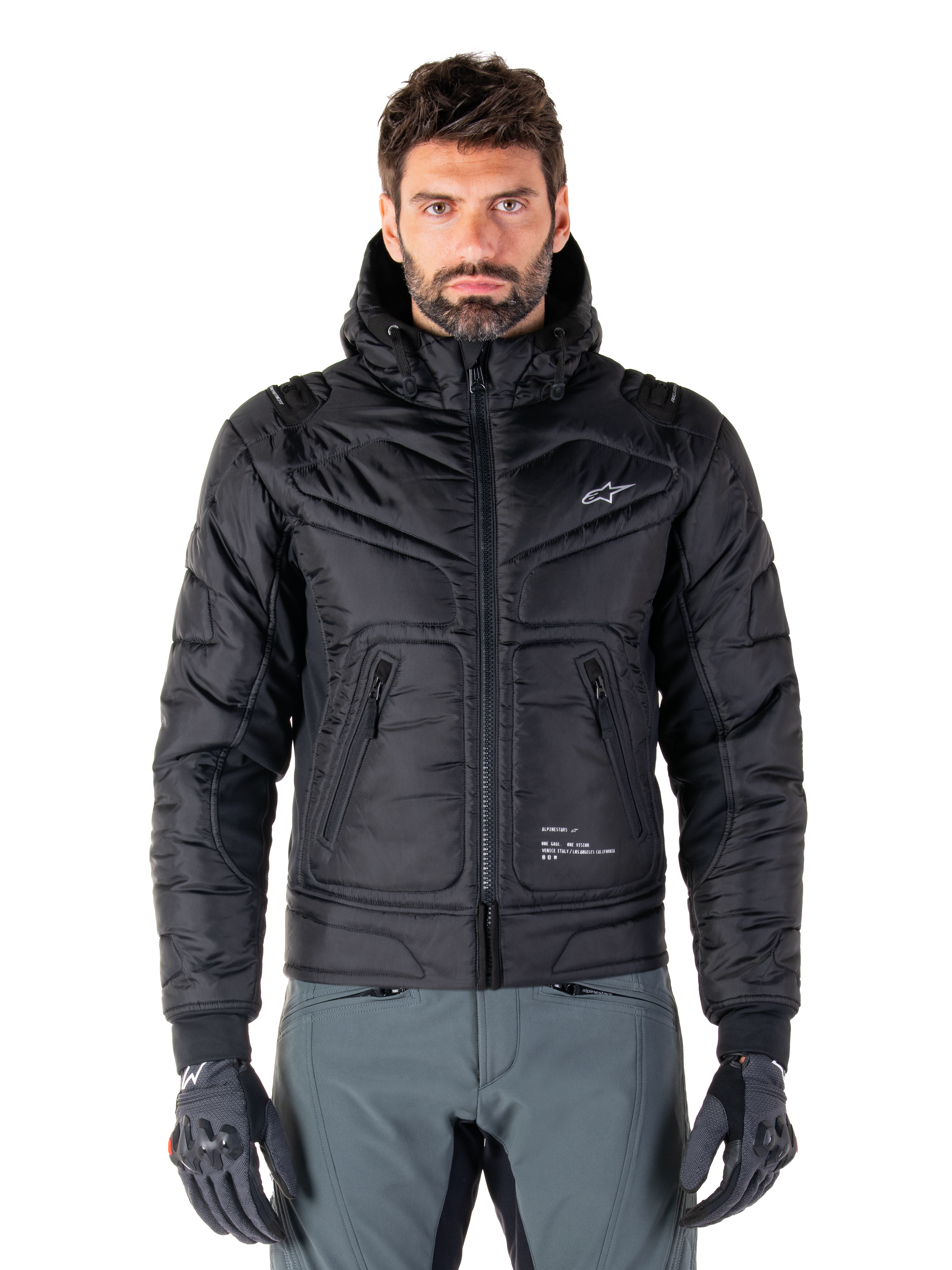 Alpinestars Mohobbs Waterproof Jacket, zwarte gewatteerde bomber motorjas, gewatteerde poly-stof met ritssluiting en ritszakken, klein wit logo op de borst, gedragen door een model met grijze broek en handschoenen, urban streetwear-ontwerp voor rijden in de stad