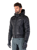 Alpinestars Mohobbs Waterproof Jacket, zwart, gewatteerd doorgestikt bombermodel met een capuchon, gedragen door een man met een grijze motorbroek en zwarte handschoenen, op urban streetwear geïnspireerde motorkleding met een klein wit logo op de borst.
