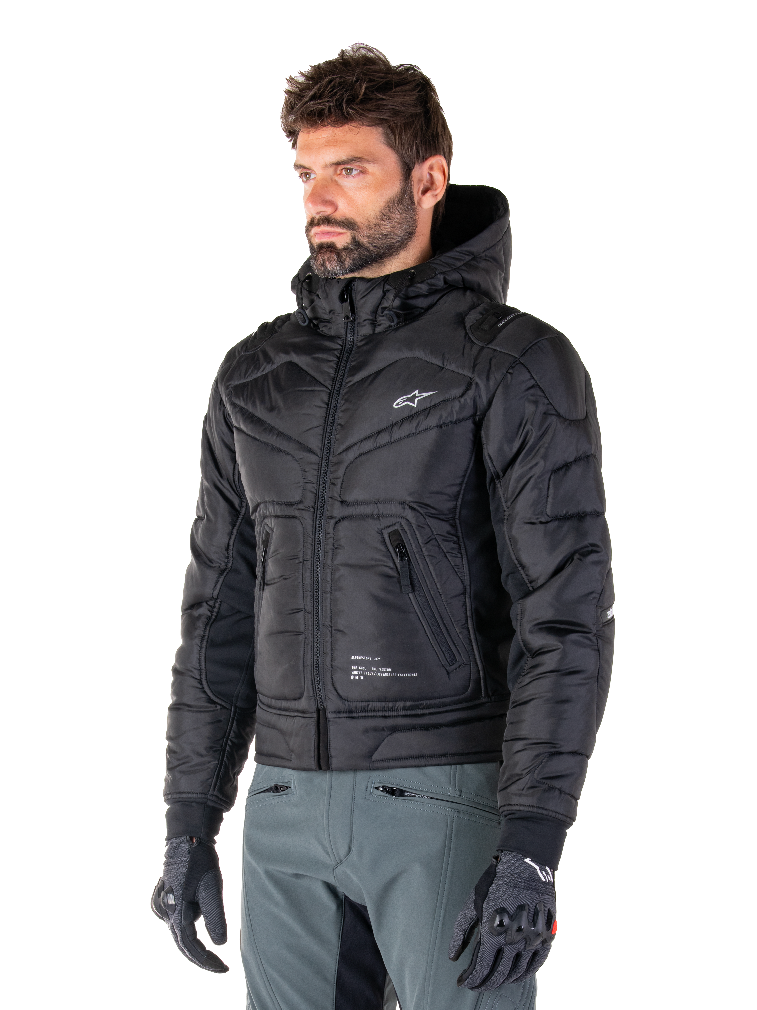 Alpinestars Mohobbs Waterproof Jacket, zwart, gewatteerd doorgestikt bombermodel met een capuchon, gedragen door een man met een grijze motorbroek en zwarte handschoenen, op urban streetwear geïnspireerde motorkleding met een klein wit logo op de borst.