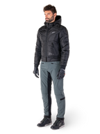 Alpinestars Mohobbs Waterproof Jacket, zwart, gewatteerde urban motorbomberjas met capuchon, gedragen door een model met grijze motorbroek en zwarte handschoenen, voorzien van een wit logo op de borst, eigentijdse op streetwear geïnspireerde beschermende kleding