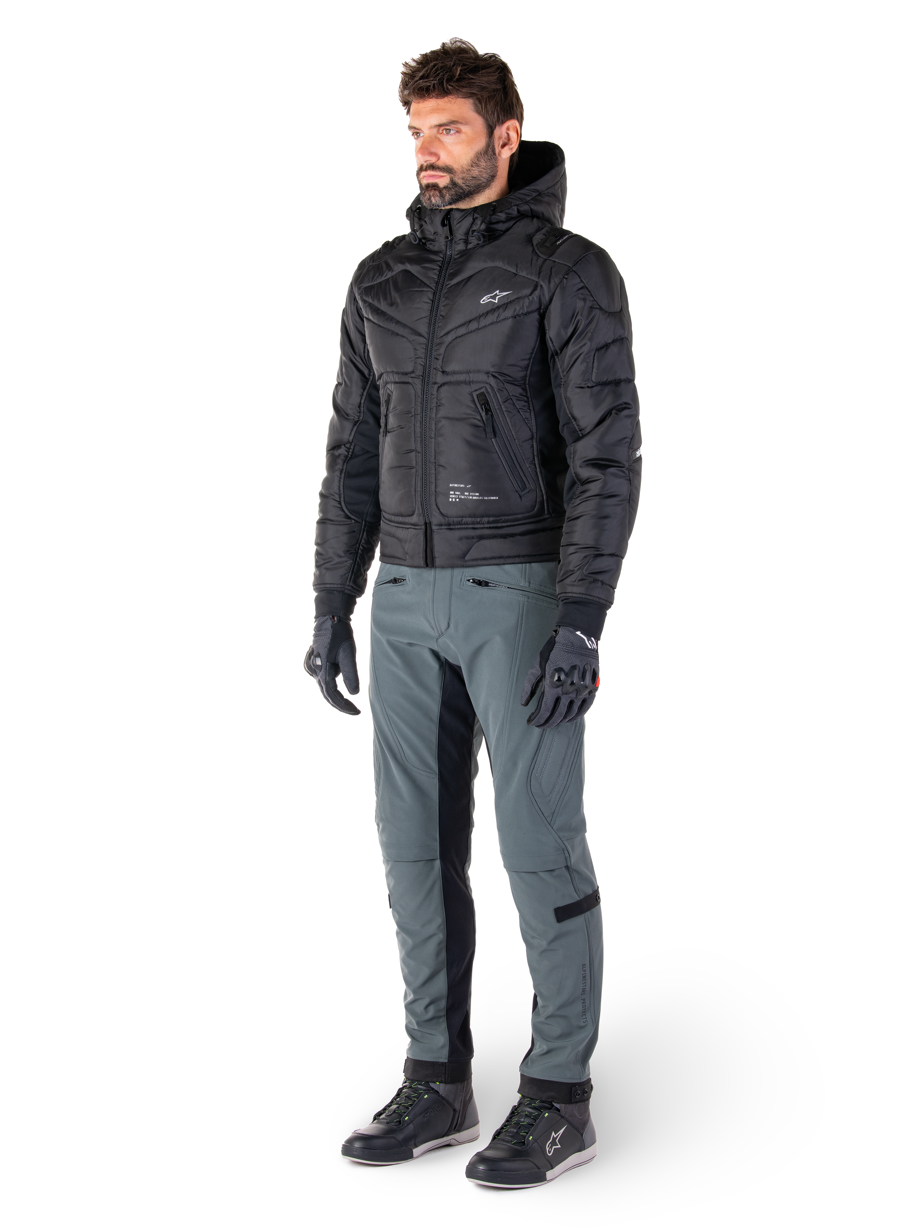 Alpinestars Mohobbs Waterproof Jacket, zwart, gewatteerde urban motorbomberjas met capuchon, gedragen door een model met grijze motorbroek en zwarte handschoenen, voorzien van een wit logo op de borst, eigentijdse op streetwear geïnspireerde beschermende kleding