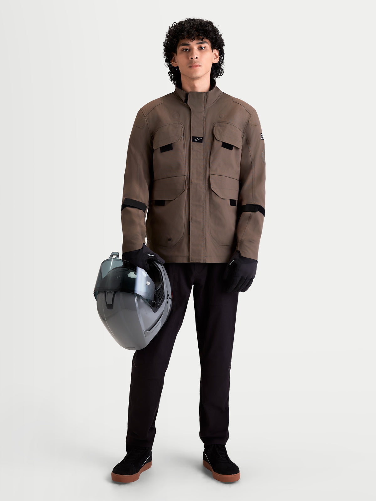 Alpinestars Altan Drystar Jacket, men's urban contemporary motorcycle utility jacket in walnut brown, front full-length view on a model wearing black pants, gloves, and holding a helmet, waterproof textile construction with cargo pockets.