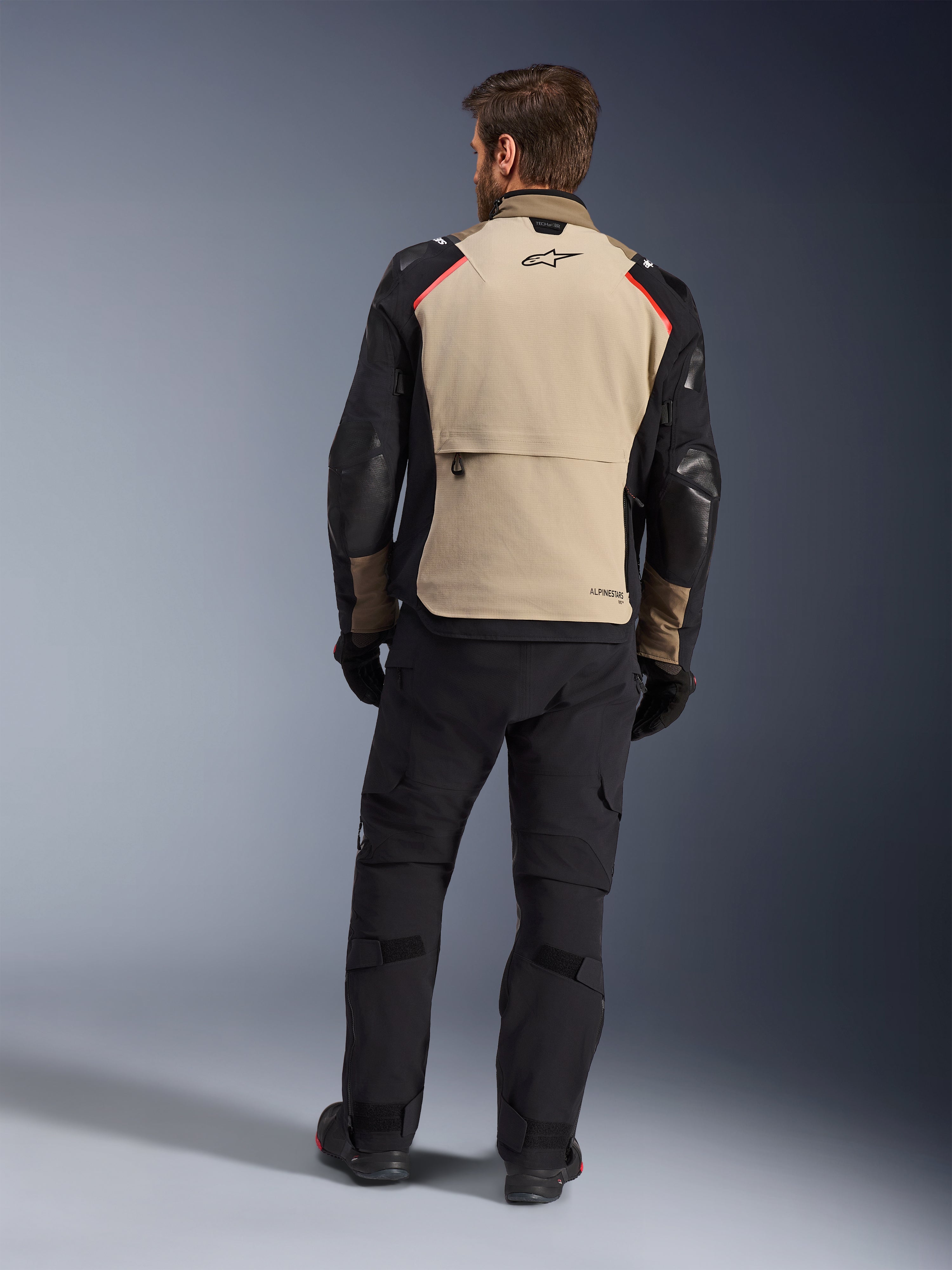 Alpinestars Andes Pro Drystar® XF Jacke, Walnut Aluminum, Rückansicht, Motorrad-Adventure-Jacke mit schwarzen Ärmeln und roten Schulterakzenten, getragen mit schwarzer Motorradhose und Handschuhen.