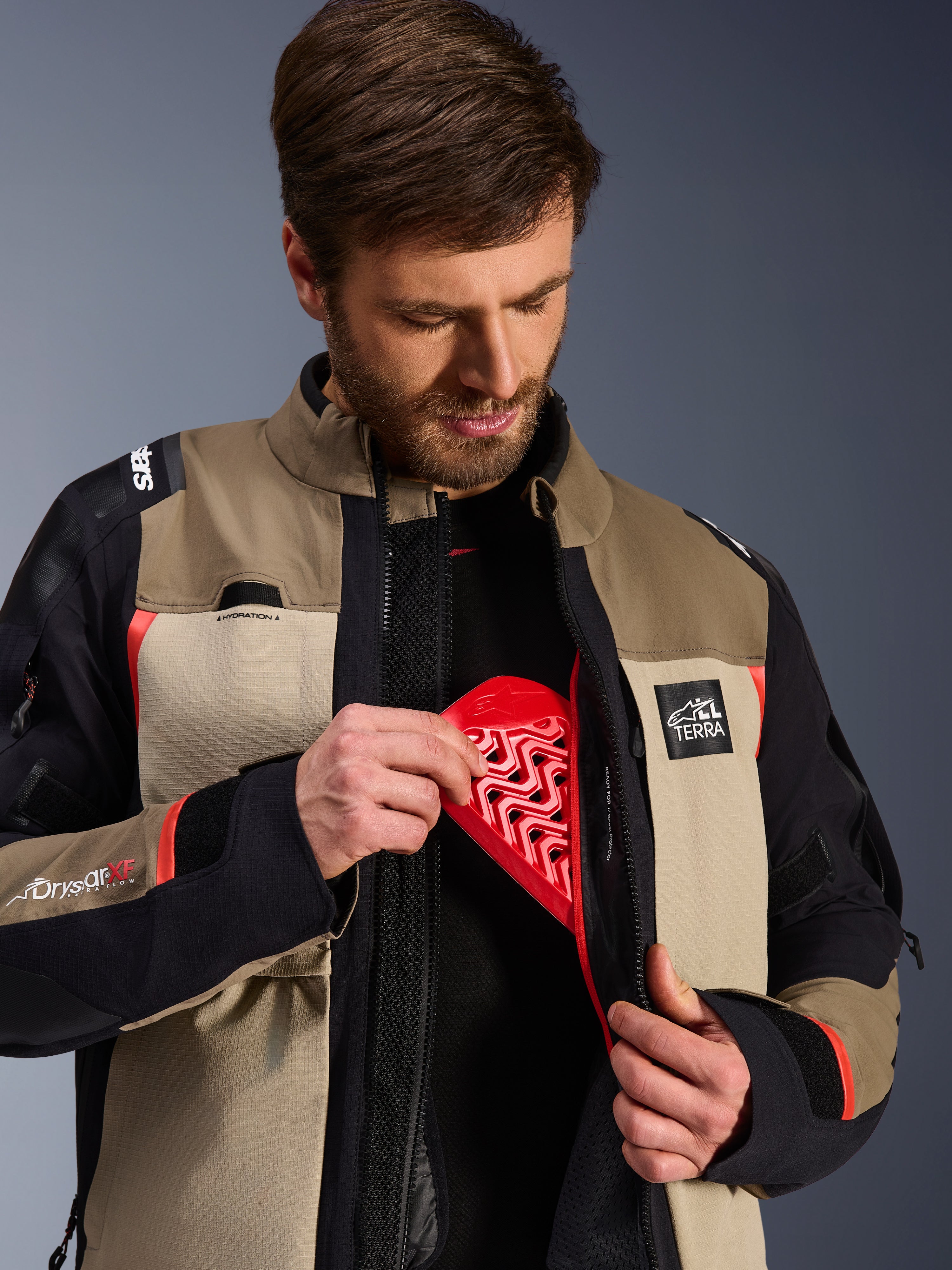 Alpinestars Andes Pro Drystar® XF Jacke in Walnut Aluminum, Modell demonstriert das Einsetzen des roten Nucleon Brustprotektors in die Innentasche, Adventure-Motorradjacke mit ALL TERRA Branding und Drystar XF Logos.