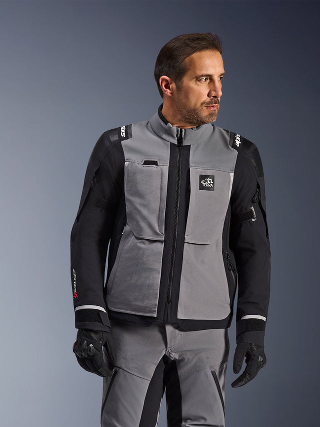 Alpinestars Andes Pro Drystar® XF jas in donkergrijs en zwart, adventure-rijuitrusting, gedragen door een man met bijpassende grijze broek en zwarte handschoenen, voorzien van een 3-laags gelamineerde buitenlaag, handige zakken en 'Terra'-logo op de borst