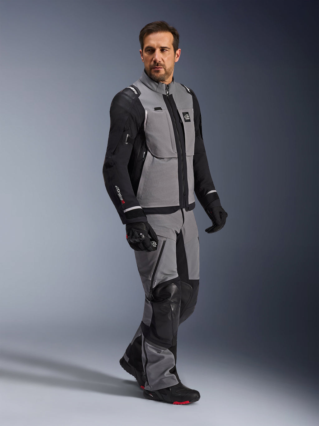 Alpinestars Andes Pro Drystar® XF Jacke, Adventure-Motorradjacke, Dunkelgrau und Schwarz, getragen von einem Modell mit passender Textilhose und schwarzen Handschuhen, mit Multi-Taschen-Design, technischem 3-Lagen-Laminat-Außenmaterial und ergonomischer Passform für Langstrecken-Adventure-Fahrten.