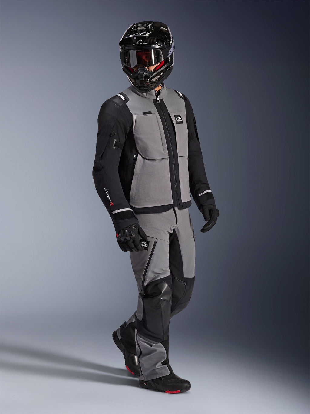 Adventure-Fahrer mit Alpinestars Andes Pro Drystar® XF Jacke in Dunkelgrau und Schwarz, dazu passende grau-schwarze Adventure-Hose, schwarzer Integralhelm mit Brille und schwarze Motorradhandschuhe, Studioaufnahme für professionelle Langstrecken-Tourenbekleidung.
