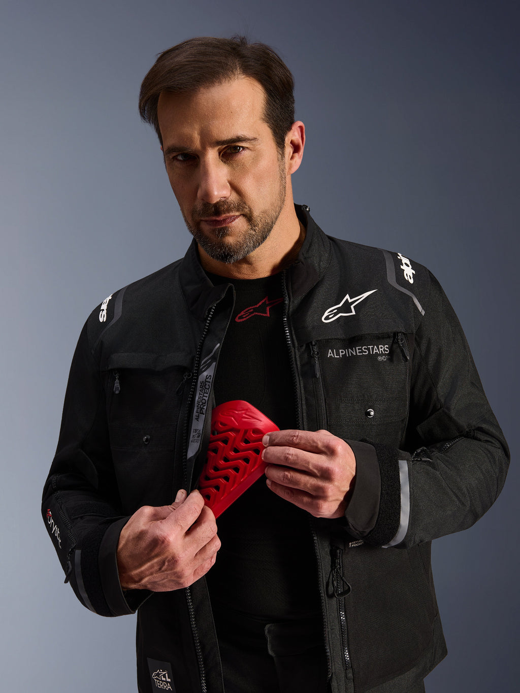 Alpinestars Cusco Drystar® Jacket in Black, male model holding a red Nucleon Flex Pro armor piece, adventure touring motorcycle jacket with white branding, technical textile construction featuring ventilation zippers and multiple pockets