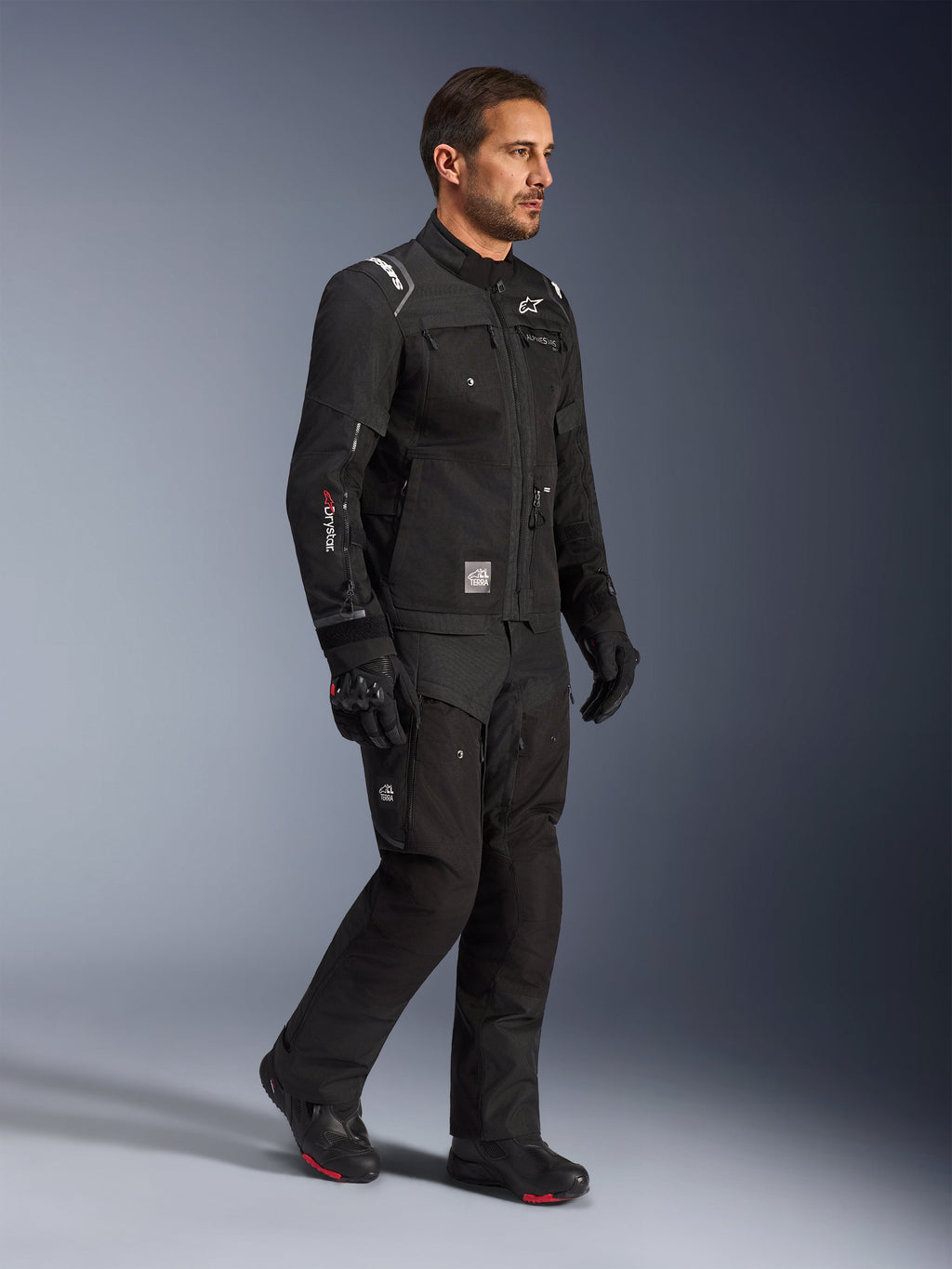 Male model wearing Alpinestars Cusco Drystar® Jacket in Black, adventure touring motorcycle gear with matching black pants and boots, featuring a multi-pocket design, 'Drystar' branding on the sleeve, and 'Terra' logo patch.