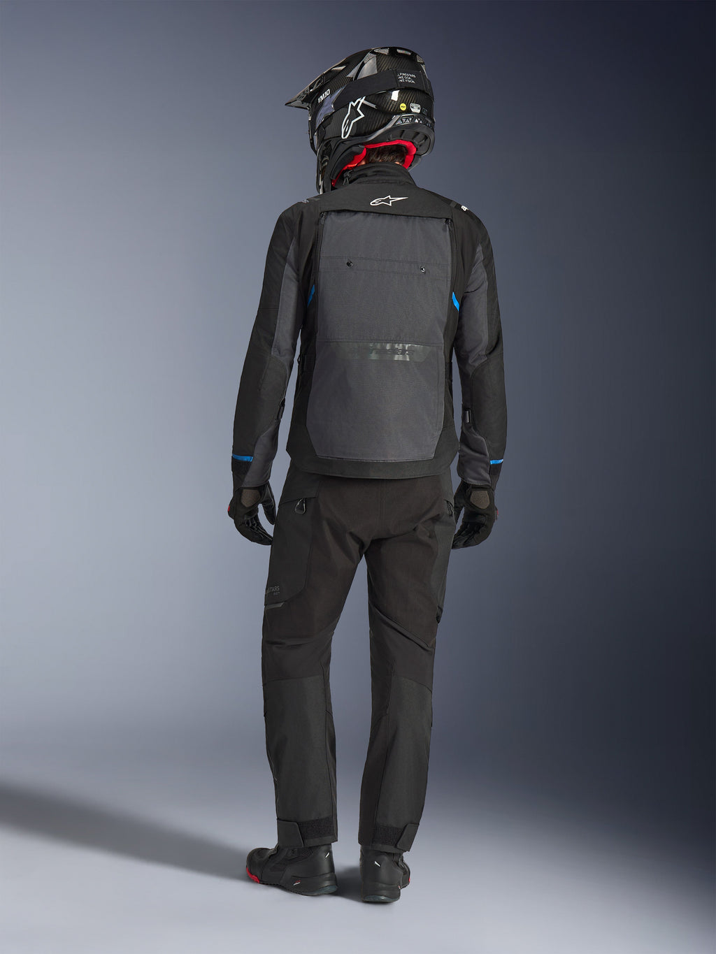 Veste Alpinestars Cusco Drystar®, noir et gris foncé, vue arrière d'un motard en tenue complète d'aventure touring, grand panneau arrière gris avec logo Alpinestars, accents bleus sur les manches, portée avec un pantalon noir et un casque assortis.