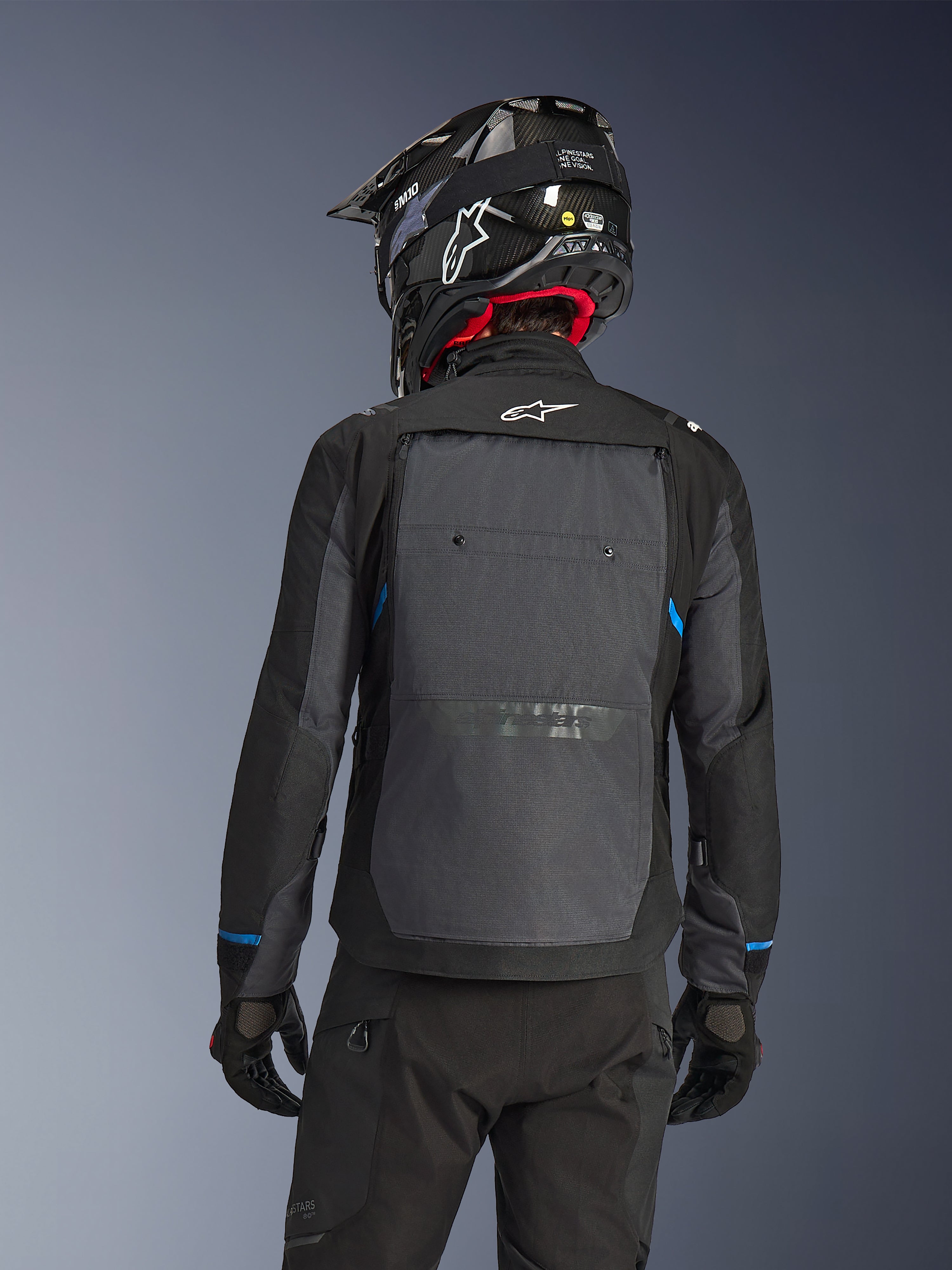 Pilote vu de dos portant la veste Alpinestars Cusco Drystar® en Noir et Gris Foncé, dotée d'un grand panneau de ventilation arrière gris et d'un logo blanc, accompagnée d'un casque tout-terrain noir et d'un équipement de moto noir pour l'adventure touring.