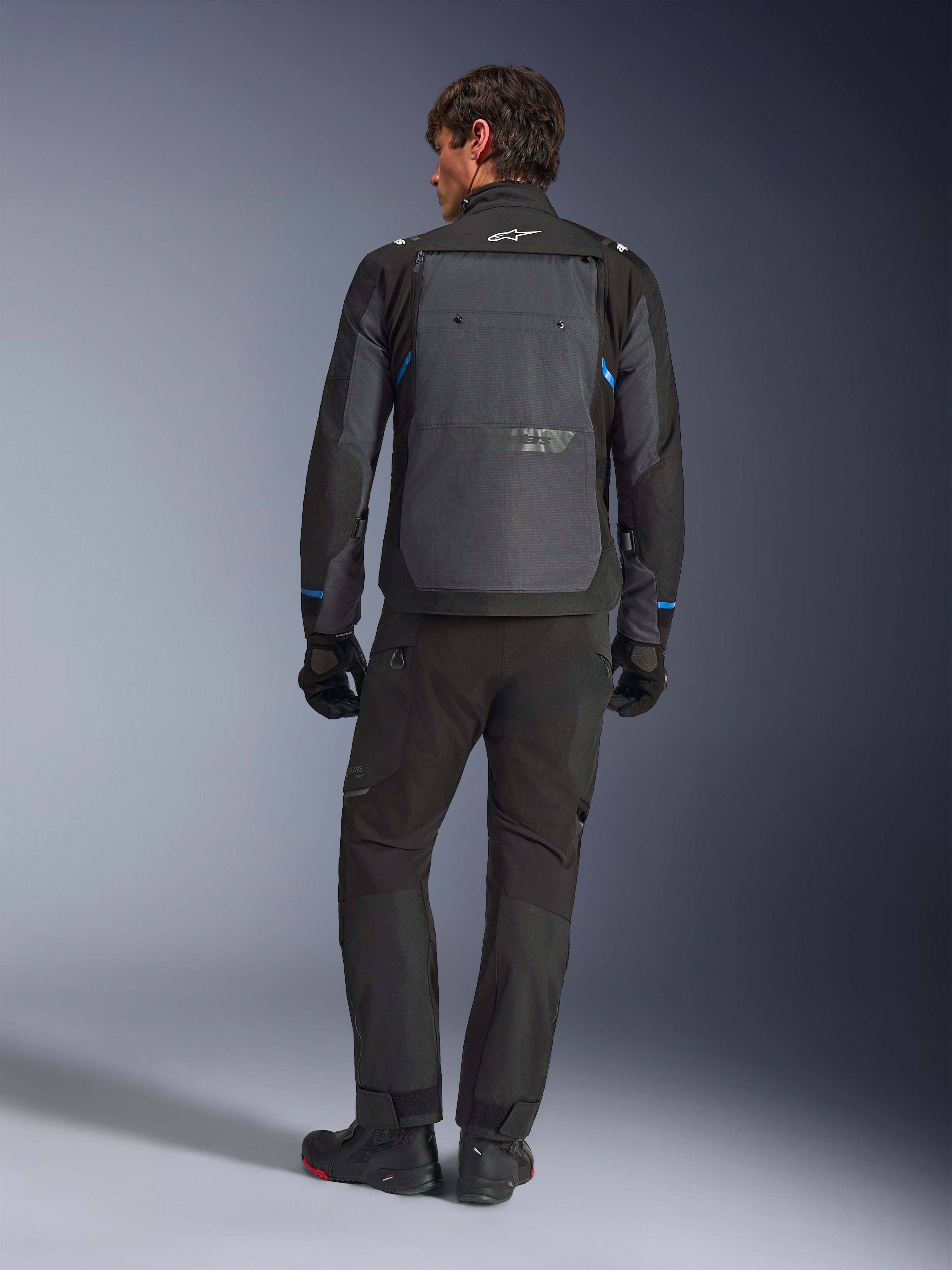 Veste Alpinestars Cusco Drystar®, noir et gris foncé avec des accents bleus, vue de dos, portée avec un pantalon d'aventure et des gants noirs assortis, dotée d'un large panneau dorsal gris et d'un logo blanc sur le col, conçue pour la route et le touring d'aventure