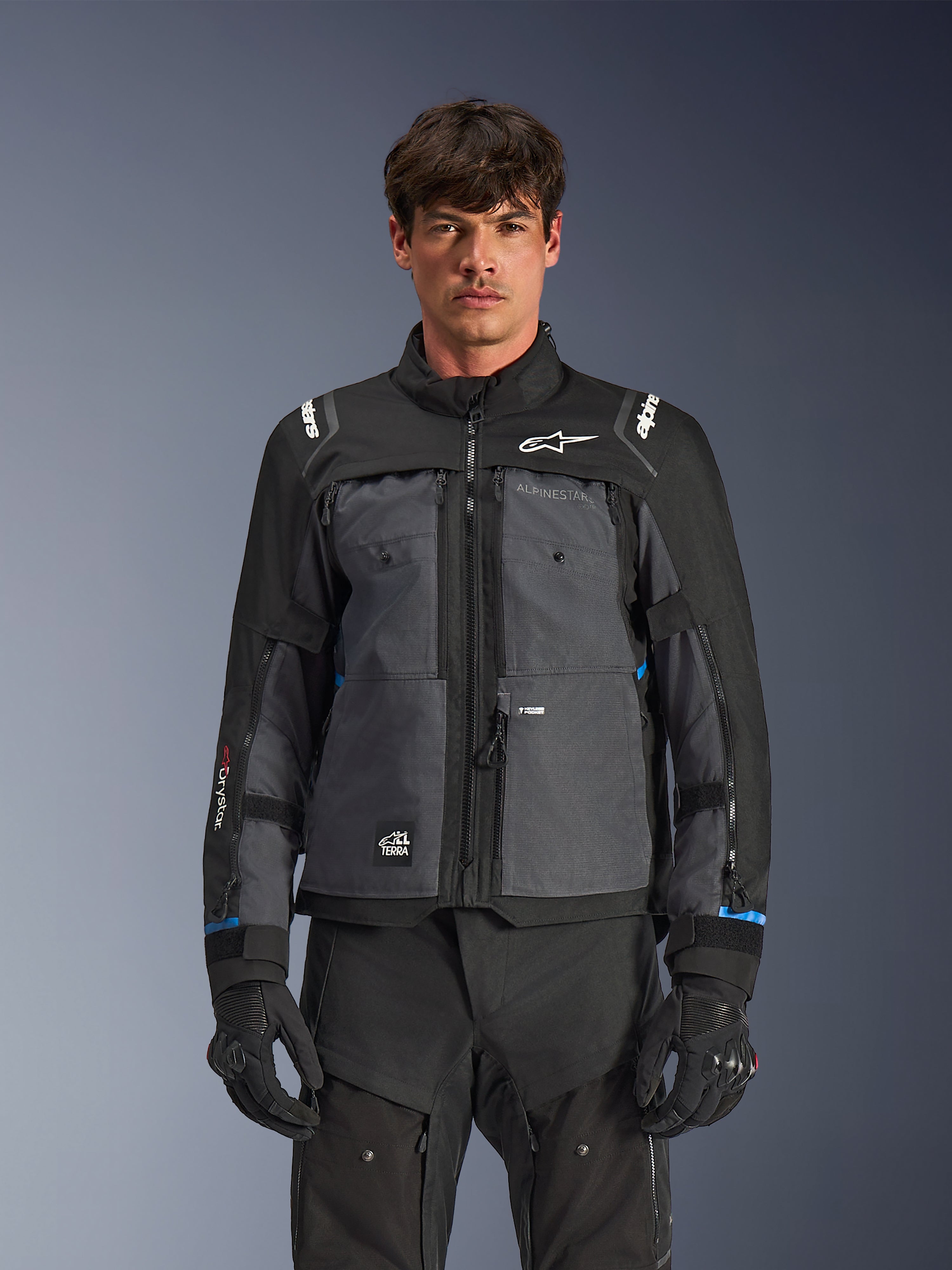 Veste Alpinestars Cusco Drystar®, veste de moto touring, noir et gris foncé, vue de face sur modèle, avec logos Alpinestars sur la poitrine et les épaules, marquage Drystar® sur la manche, design technique multi-poches avec zips de ventilation, associée à un équipement de moto noir