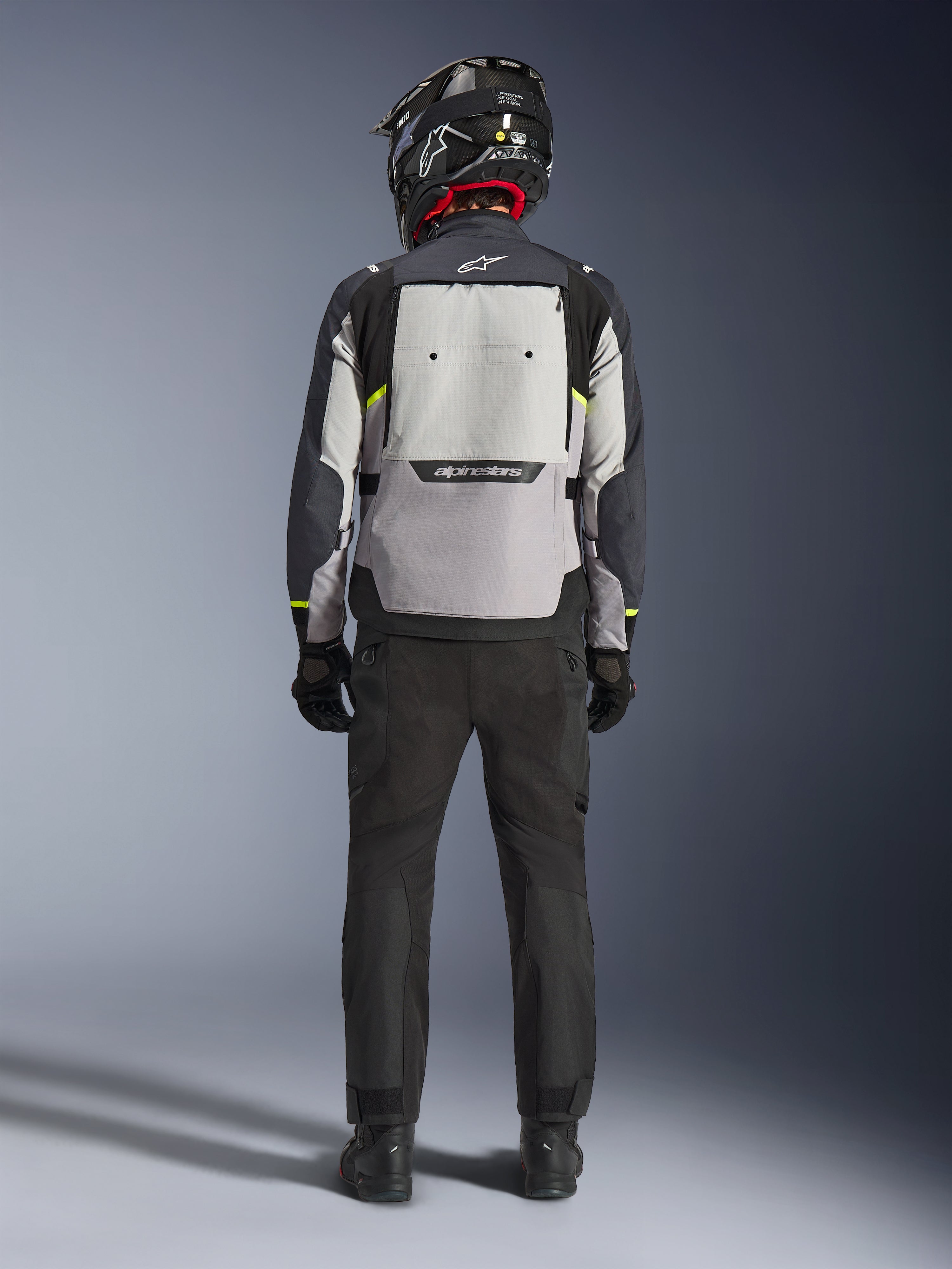 Motard vu de dos portant la veste Alpinestars Cusco Drystar® en Ice Gray, Mid Gray et Deep Gray, dotée d'un large panneau de ventilation dorsal et du logo Alpinestars, avec un pantalon de moto noir et un casque noir, équipement de moto adventure touring