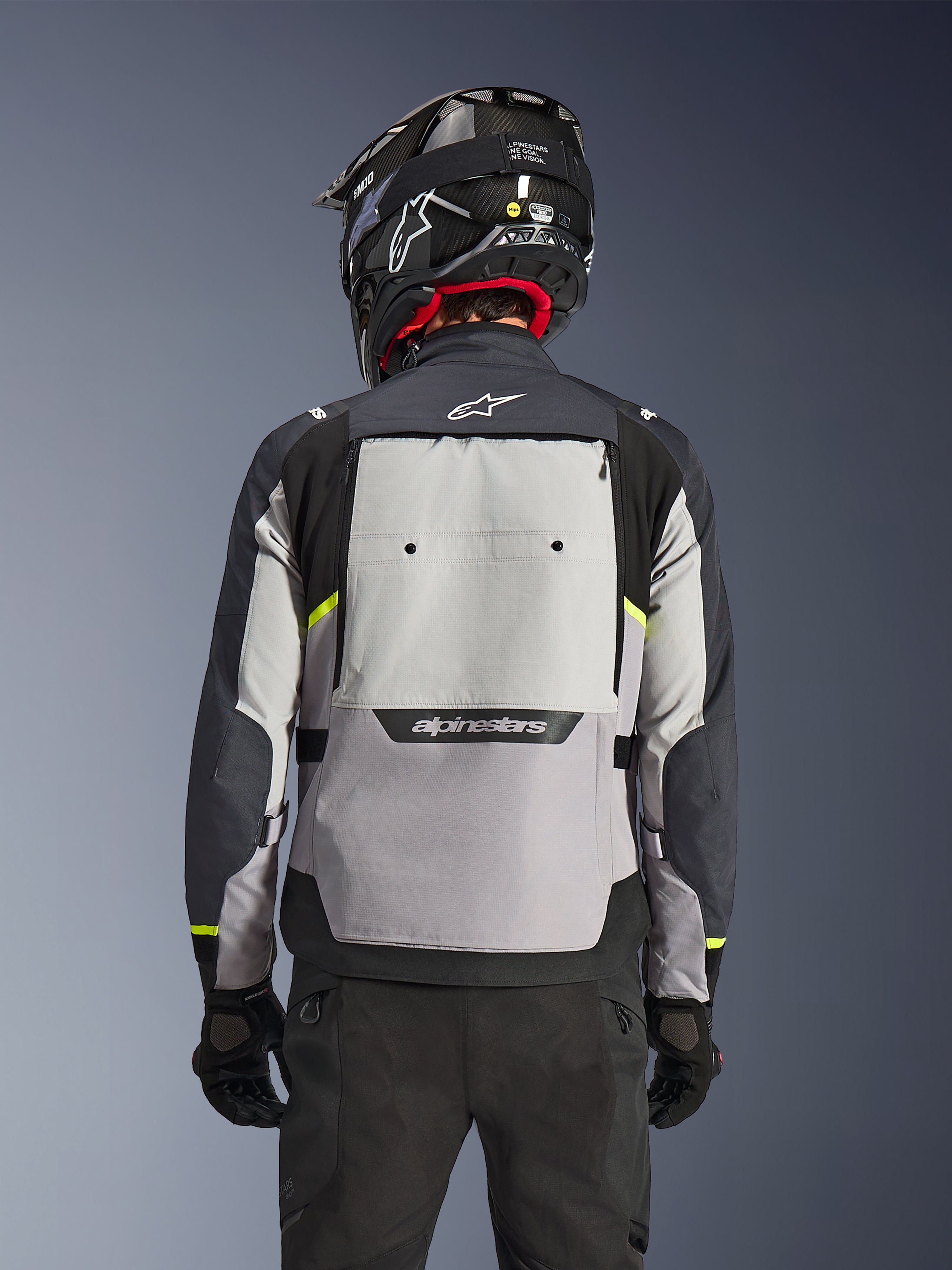 Veste Alpinestars Cusco Drystar®, vue arrière, coloris Ice Gray, Mid Gray et Deep Gray, dotée d'un large panneau de ventilation dorsal et d'accents jaune haute visibilité, portée avec un casque d'aventure noir et un pantalon noir.