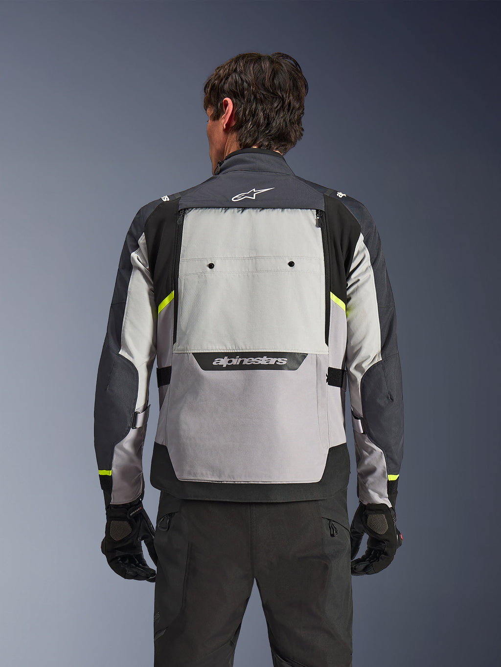 Vue arrière d'un motard portant la veste Alpinestars Cusco Drystar® en Gris Glace, Gris Moyen et Gris Foncé, présentant des accents jaune néon, un large panneau de ventilation dorsal et un marquage Alpinestars audacieux, associée à un pantalon et des gants de moto noirs.