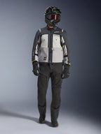 Veste Alpinestars Cusco Drystar®, Gris Glace, Gris Moyen et Gris Foncé, veste de moto d'aventure portée avec un pantalon de moto noir, des gants noirs et un casque noir avec lunettes, dotée de panneaux gris contrastés et de détails techniques pour la route et le touring d'aventure.