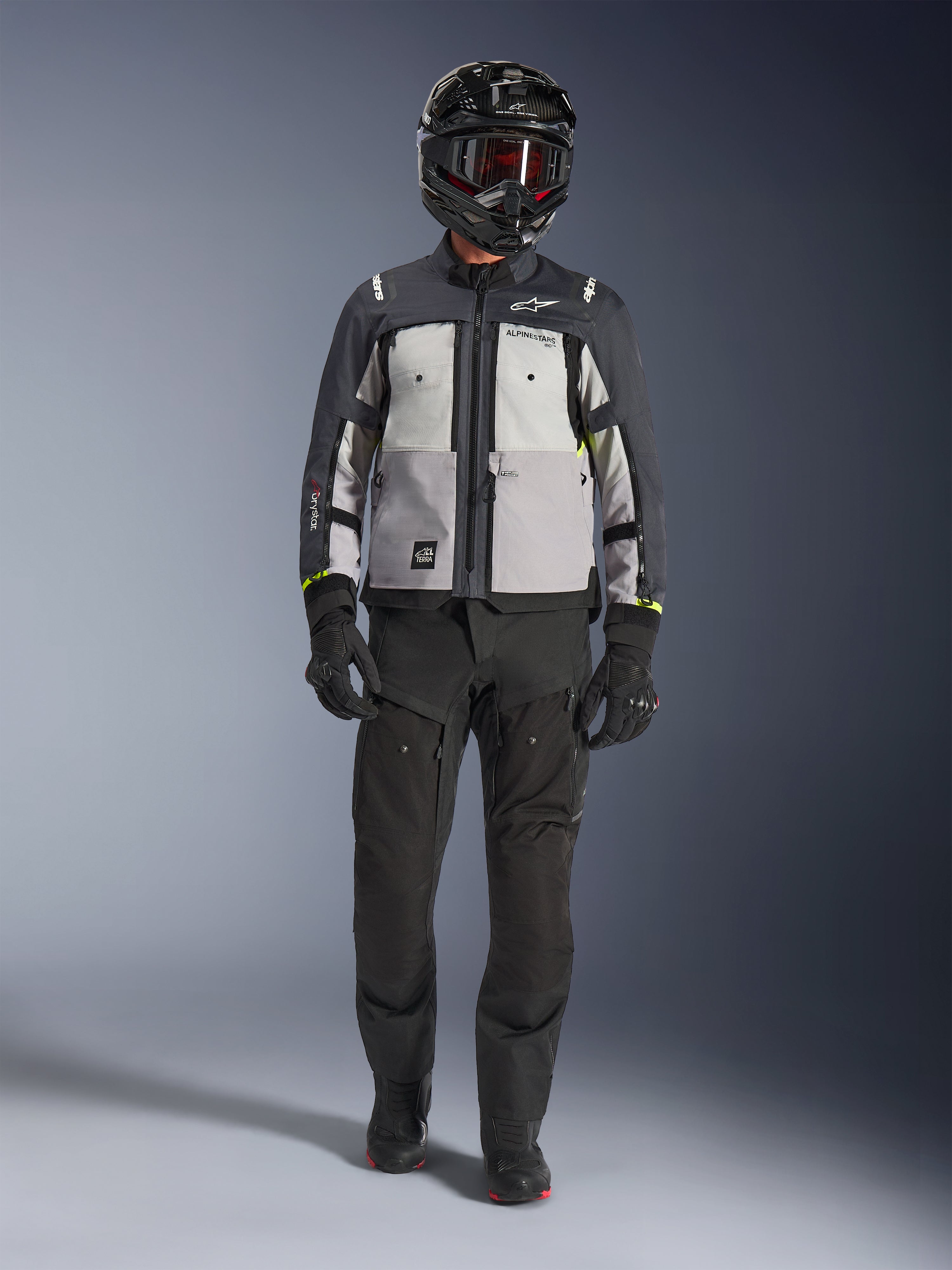 Veste Alpinestars Cusco Drystar®, Gris Glace, Gris Moyen et Gris Foncé, veste de moto d'aventure portée avec un pantalon de moto noir, des gants noirs et un casque noir avec lunettes, dotée de panneaux gris contrastés et de détails techniques pour la route et le touring d'aventure.