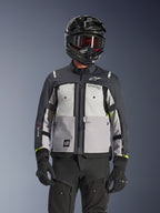 Veste Alpinestars Cusco Drystar®, veste d'aventure touring, coloris Ice Gray Mid Gray et Deep Gray, dotée de panneaux gris multi-tons, de ventilations zippées sur la poitrine et du marquage Alpinestars, portée par un pilote avec un casque et un masque noirs.