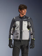 Veste Alpinestars Cusco Drystar®, veste de moto adventure touring, gris glace, gris moyen et gris foncé, portée par un mannequin avec un pantalon et des gants noirs, présentant un design textile technique multi-panneaux, des poches de poitrine, le marquage « Drystar » sur la manche et des touches discrètes de jaune fluo aux poignets.