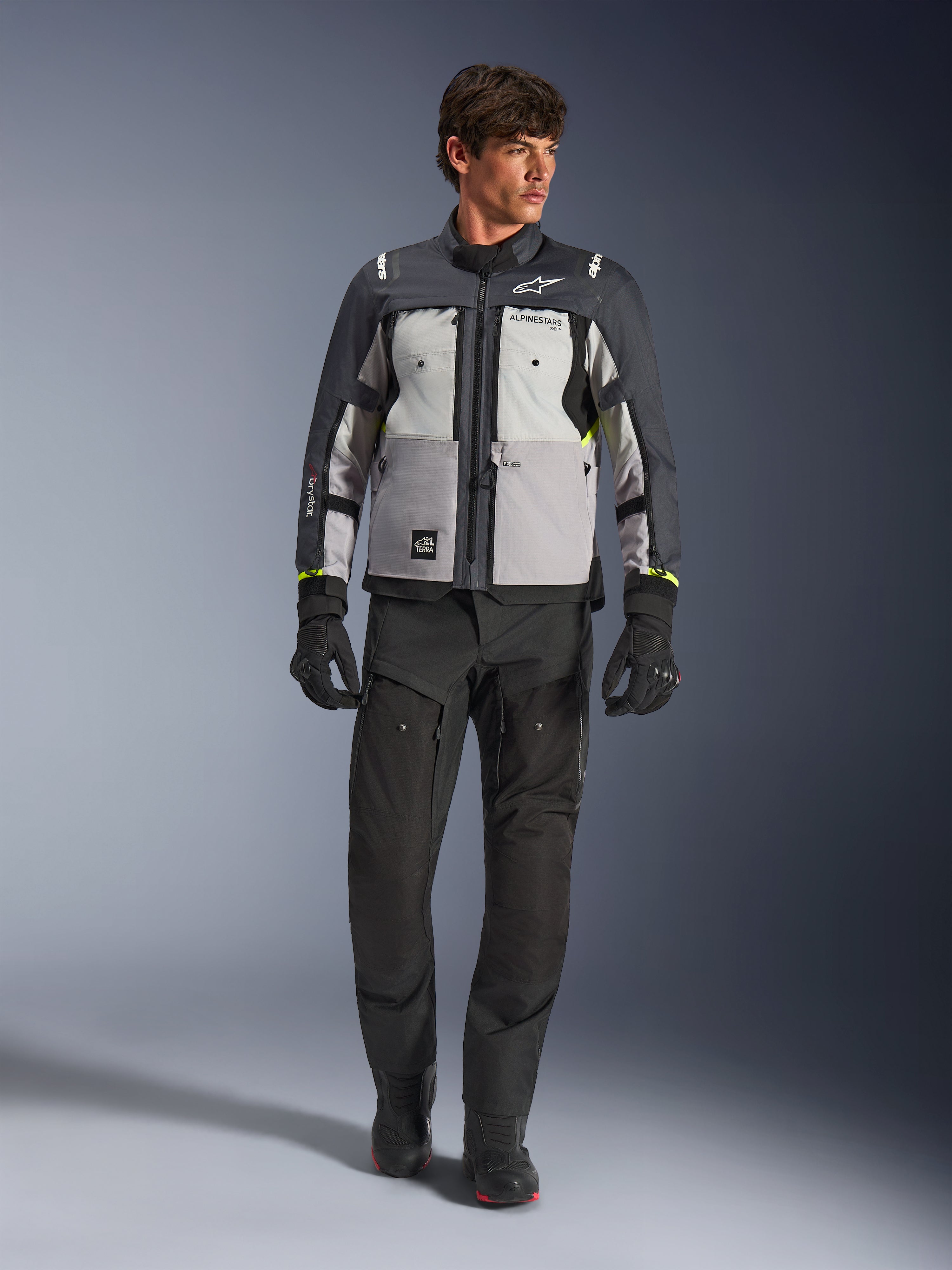 Veste Alpinestars Cusco Drystar® en Ice Gray, Mid Gray et Deep Gray, portée avec un pantalon et des gants de moto adventure noirs, veste de touring adventure en textile avec panneaux gris multi-tons et accents jaune fluo, photo de studio d'un mannequin homme.