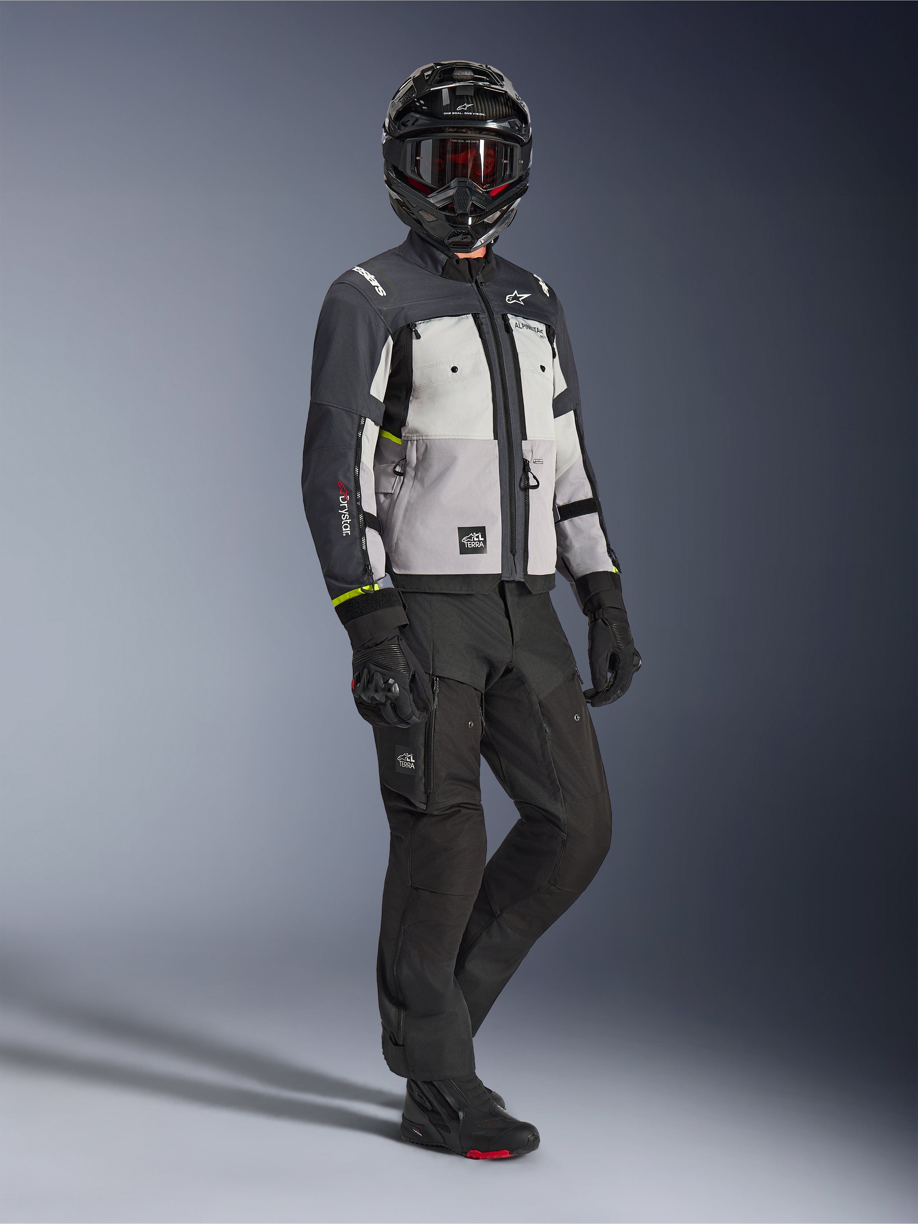 Veste Alpinestars Cusco Drystar® en Ice Gray Mid Gray Deep Gray, vue en pied d'un motard portant un équipement d'aventure complet comprenant un casque noir avec des lunettes rouges, un pantalon noir assorti et des gants, veste de moto en textile avec panneaux de ventilation directe et membrane imperméable.