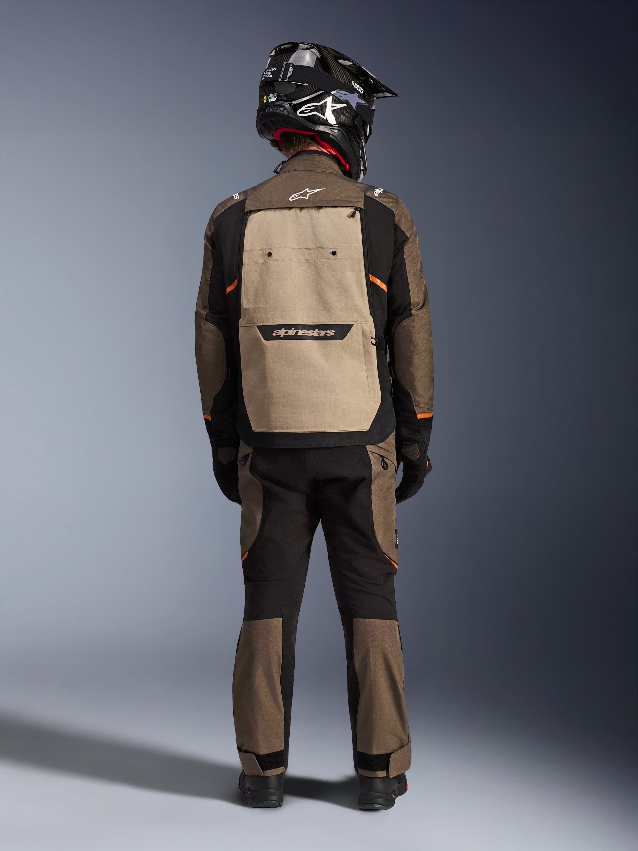 Rider from the back wearing the Alpinestars Cusco Drystar® Jacket in Walnut Aluminum Black, featuring a large aluminum-colored back pocket and orange accents, matching adventure pants, black off-road helmet, and gloves, durable textile motorcycle gear for touring.
