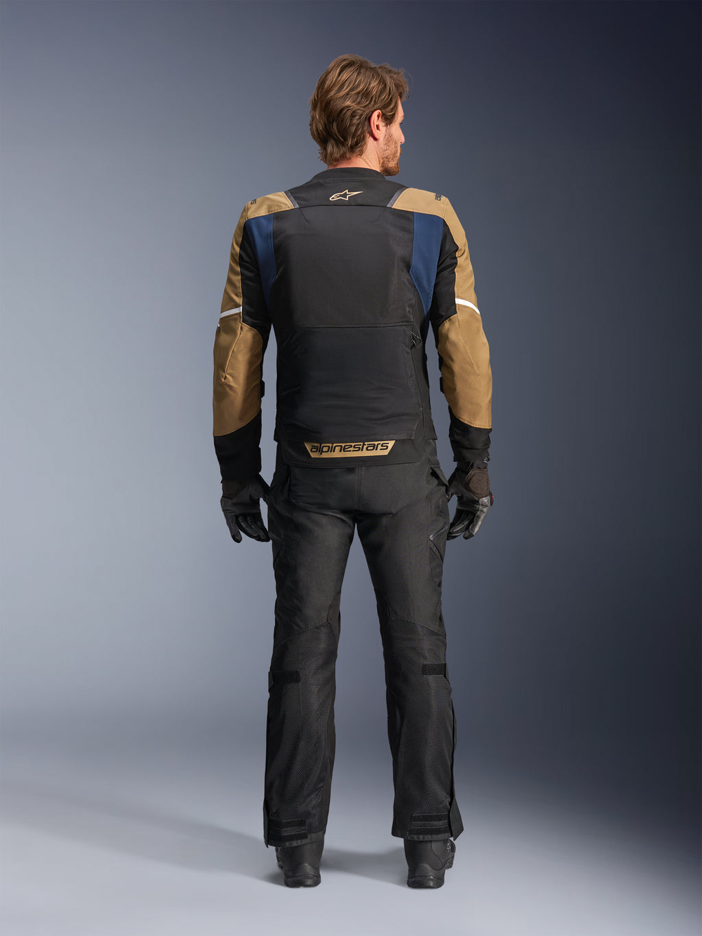 Alpinestars ST-2 Air Jacket in Deep Blue, Dark Khaki, and Black, rear view on model showing large mesh back panel and khaki sleeves with white stripes, paired with black technical motorcycle pants and gloves for adventure touring.