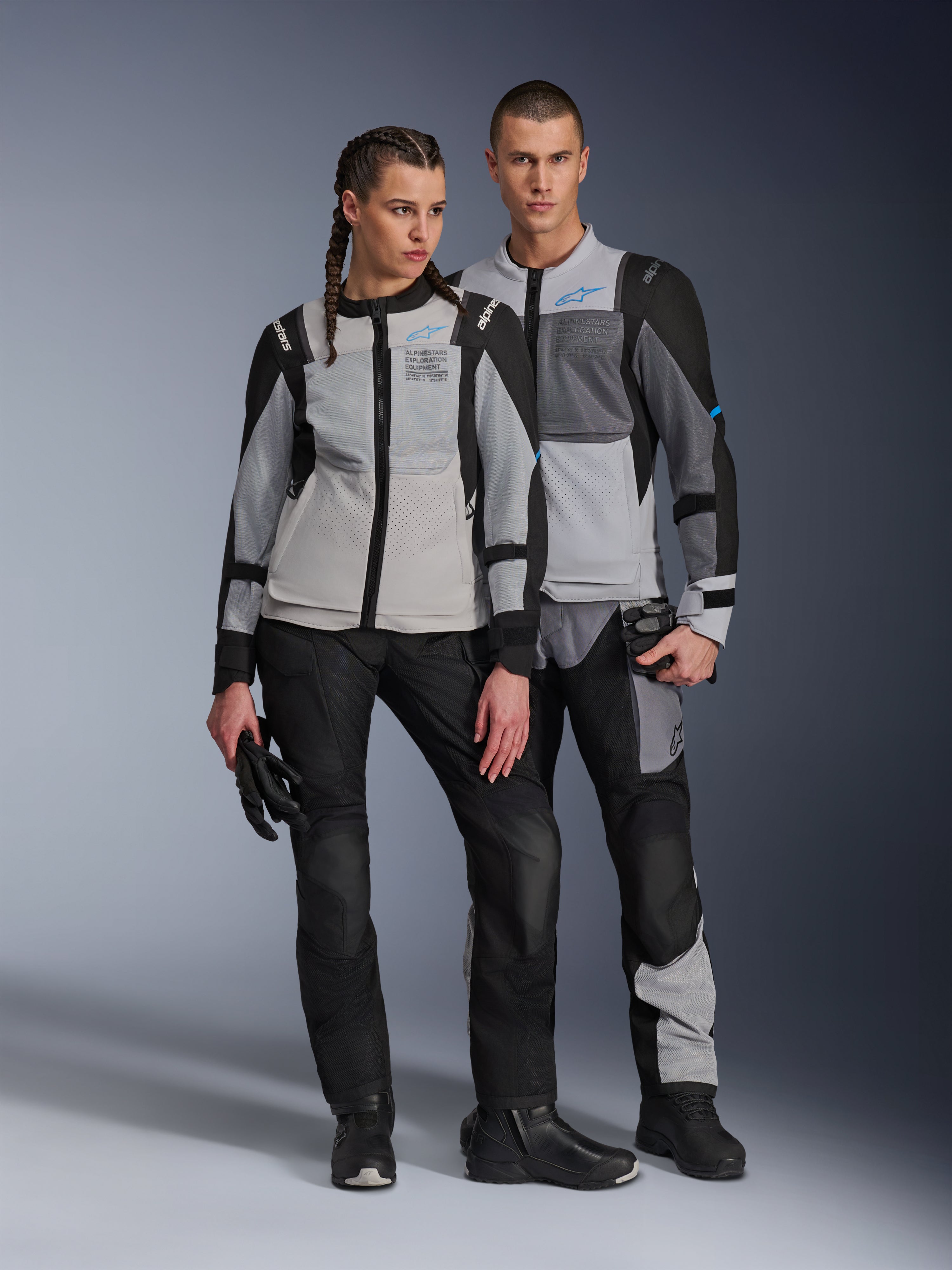 Männliches und weibliches Model in Alpinestars ST-2 Air Jacken in Ice Gray, Dark Gray und Schwarz, technische Sommer-Touring-Motorradausrüstung mit großen Mesh-Einsätzen und Exploration Equipment-Branding, kombiniert mit schwarzen Motorradhosen, Handschuhen und Stiefeln.