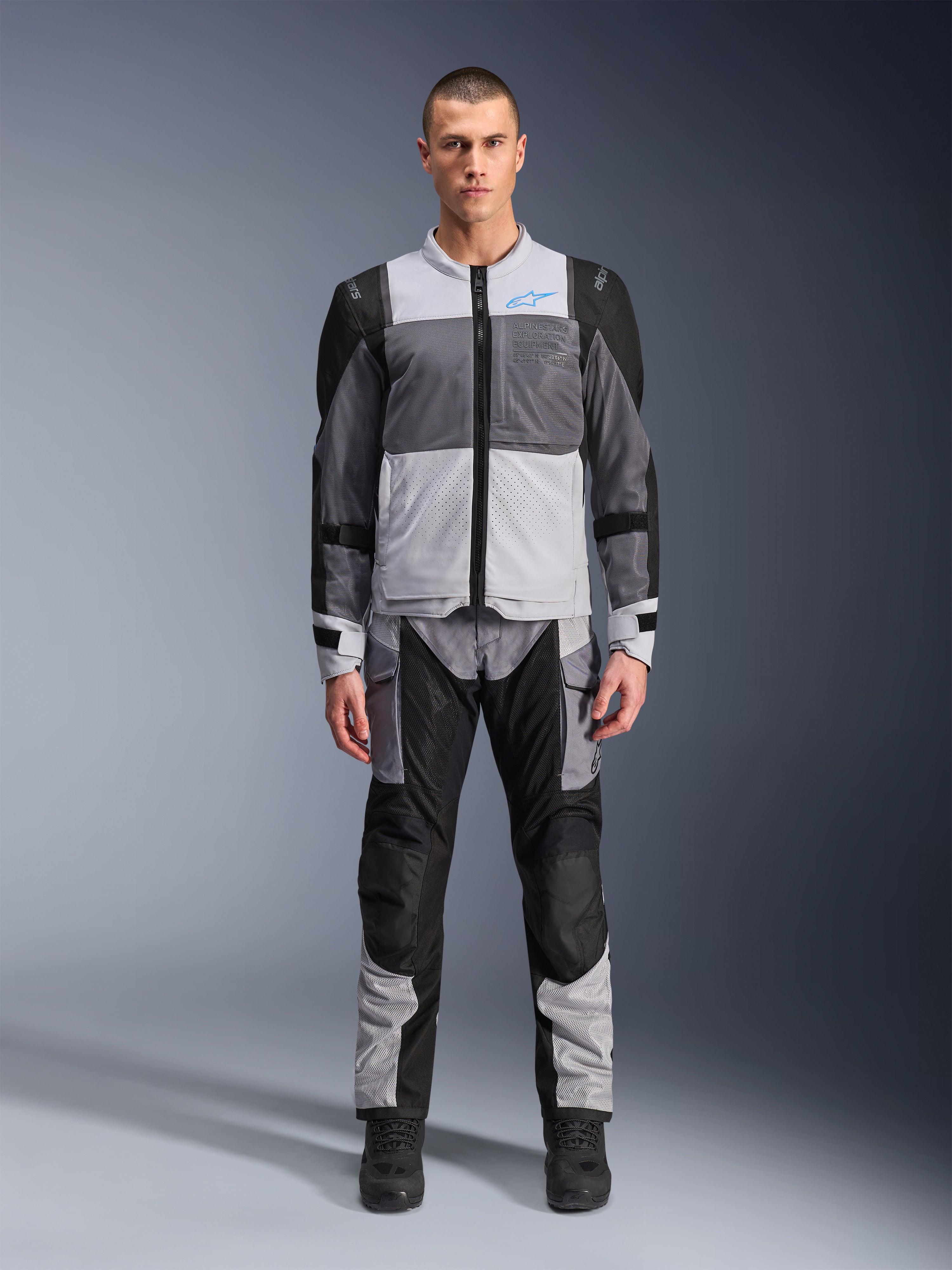 Alpinestars ST-2 Air Jacke, Motorrad-Tourenjacke, Eisgrau, Dunkelgrau und Schwarz, getragen von einem Modell mit passender Adventure-Hose, mit großen Mesh-Einsätzen, blauem Alpinestars-Logo auf der Brust und Alpinestars Exploration Equipment-Branding.